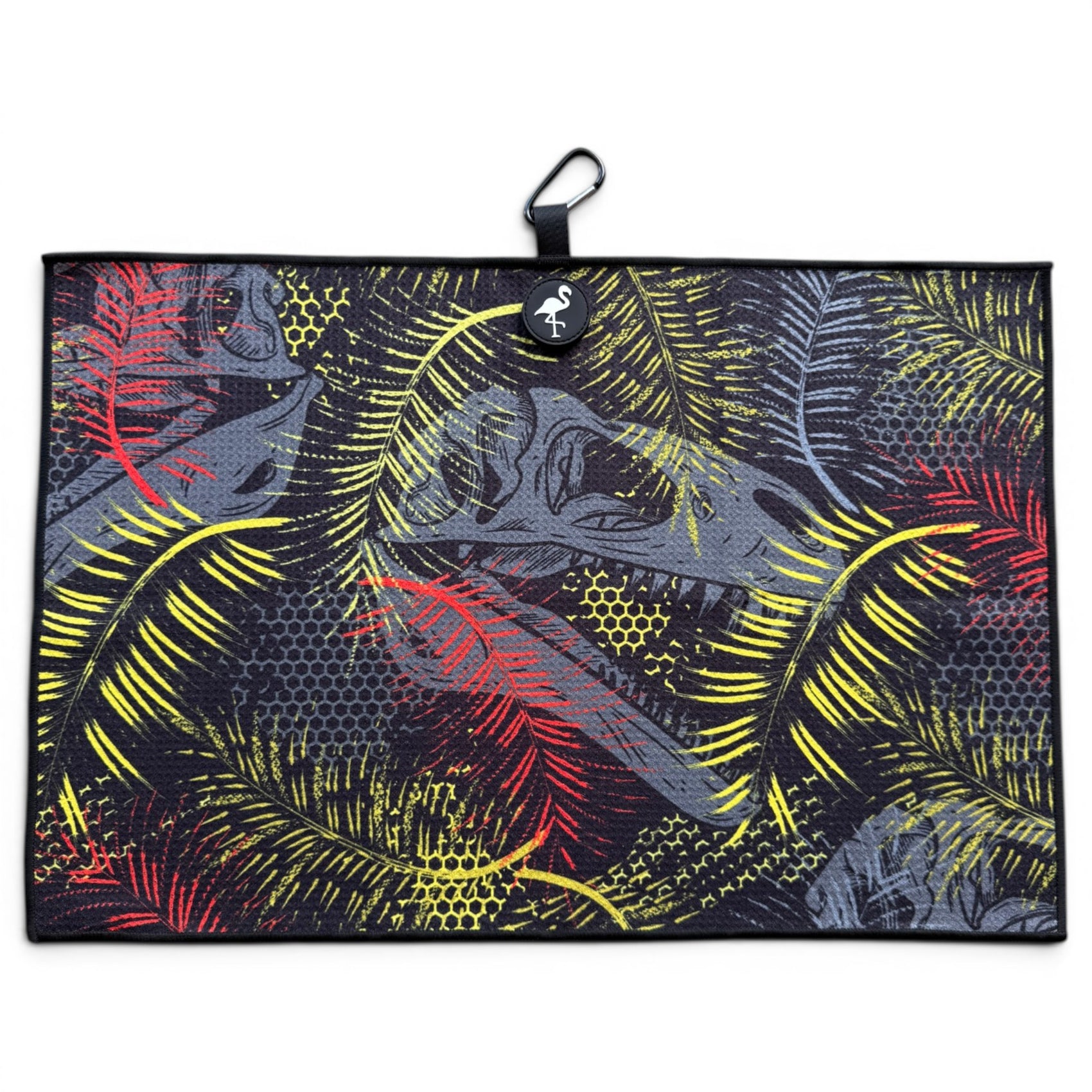 The Jurassic Fairway Golf Towel (24” x 16”) is a rectangular microfiber towel with a black background, red/yellow/gray tropical leaves, black border, hanging loop, white animal logo tag at top center, plus magnetic clip and carabiner.