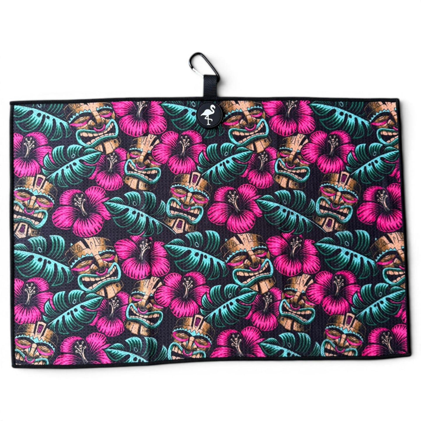 The Tiki Totem Golf Towel is a 24” x 16” microfiber towel with a vibrant tropical design of pink hibiscus, green leaves, and tan tiki masks on black. Features a magnetic clip, carabiner, and hanging loop for easy attachment.