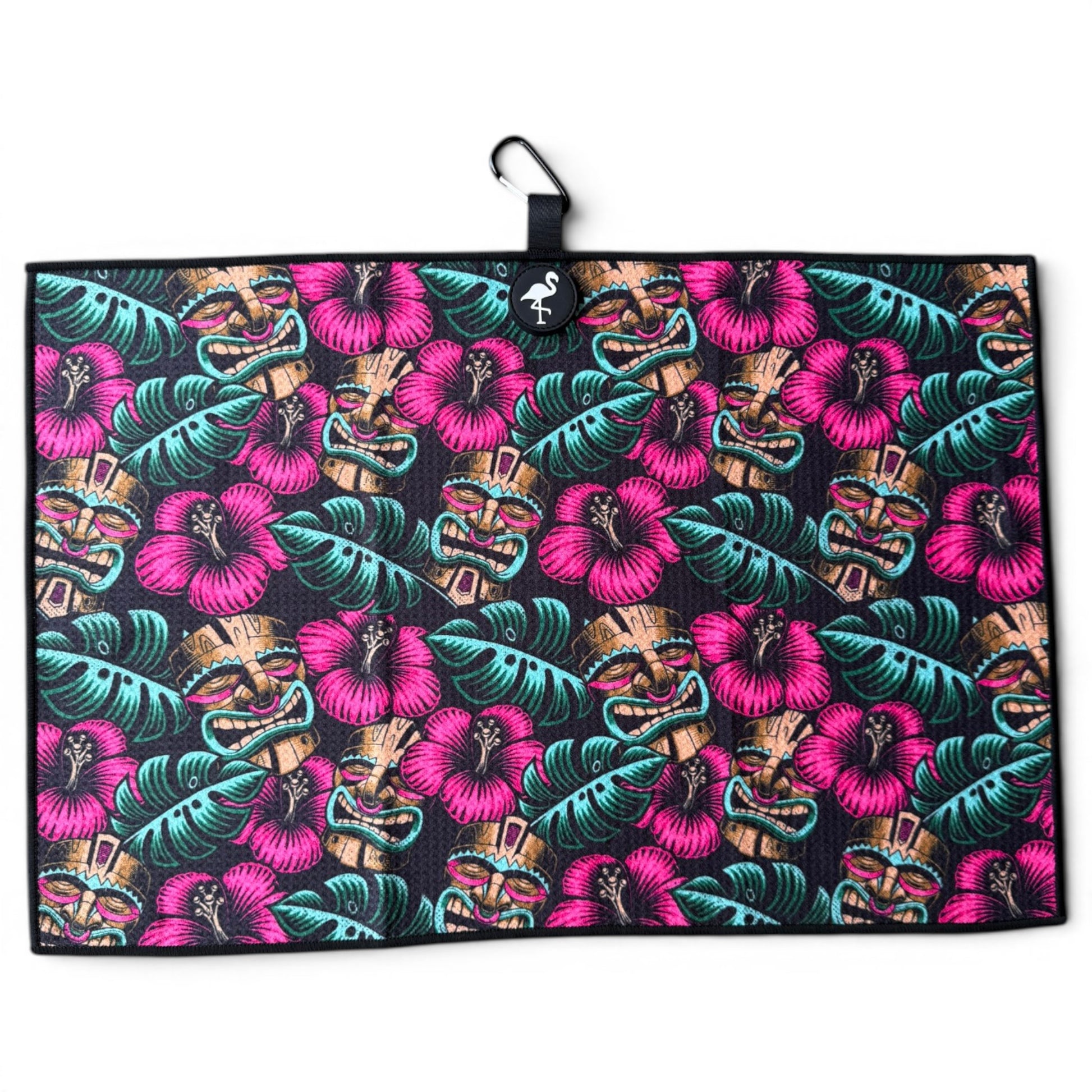 The Tiki Totem Golf Towel is a 24” x 16” microfiber towel with a vibrant tropical design of pink hibiscus, green leaves, and tan tiki masks on black. Features a magnetic clip, carabiner, and hanging loop for easy attachment.