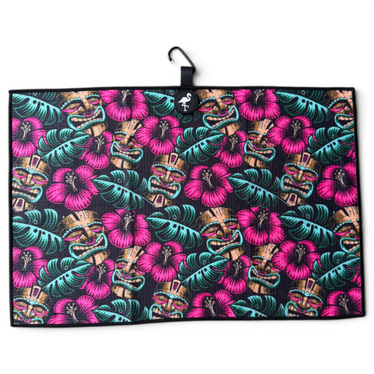 The Tiki Totem Golf Towel is a 24” x 16” microfiber towel with a vibrant tropical design of pink hibiscus, green leaves, and tan tiki masks on black. Features a magnetic clip, carabiner, and hanging loop for easy attachment.