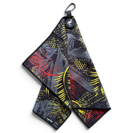 The Jurassic Fairway Golf Towel is a 24” x 16” microfiber towel with abstract red and yellow patterns on a black base, a flamingo logo tag, and includes both a magnetic clip and carabiner for easy hanging.