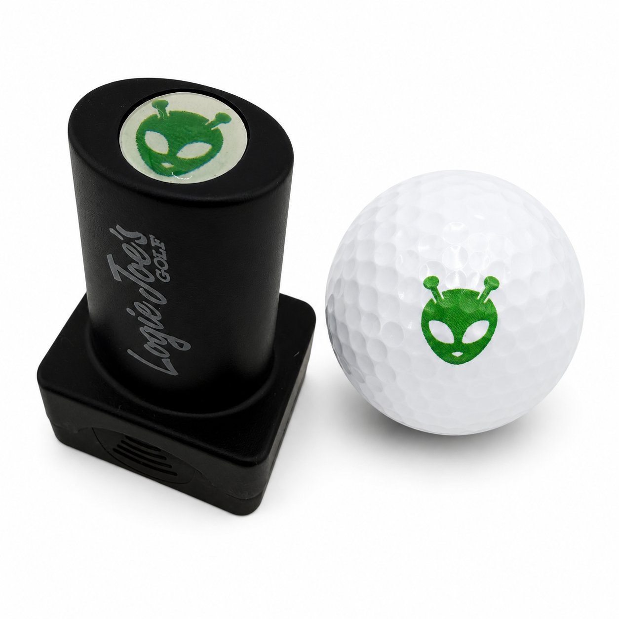 The Alien Golf Ball Stamp, featuring quick-dry ink and a green alien face logo, sits beside a white golf ball—making ball identification easy and adding fun to your game with this unique personalized accessory.