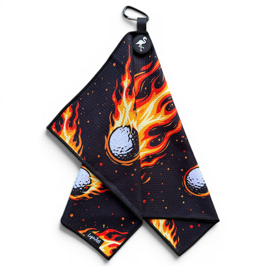 The Fireball Drive Golf Towel (24” x 16”) features black microfiber with a red, orange, and yellow flaming golf ball pattern, magnetic clip, carabiner, and round tag with flamingo logo—a premium golf accessory.