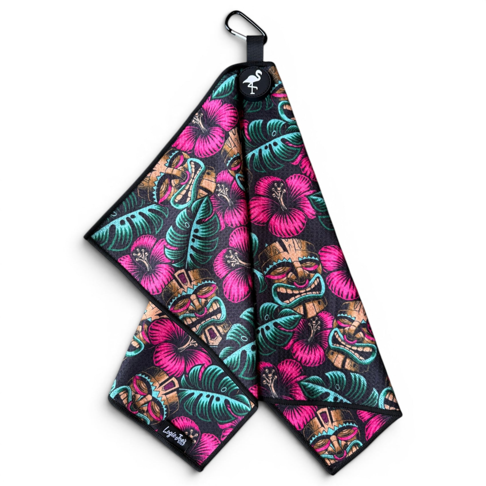 The Tiki Totem Golf Towel (24”x16”) features a tropical design with pink hibiscus, green leaves, and tiki faces. This premium microfiber towel has a magnetic clip, carabiner, and flamingo logo—perfect for keeping your golf gear stylishly clean.