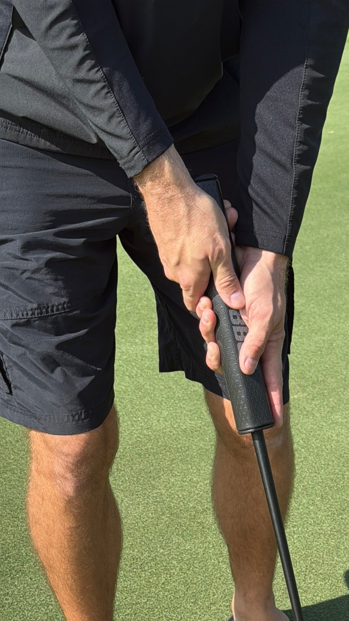A person grips The Big Black Grip "The BBG" 5.0 Tour Oversized Putter Grip, made from premium PU for maximum stability, while preparing to putt on a golf green. Only their hands and legs in black attire are visible.
