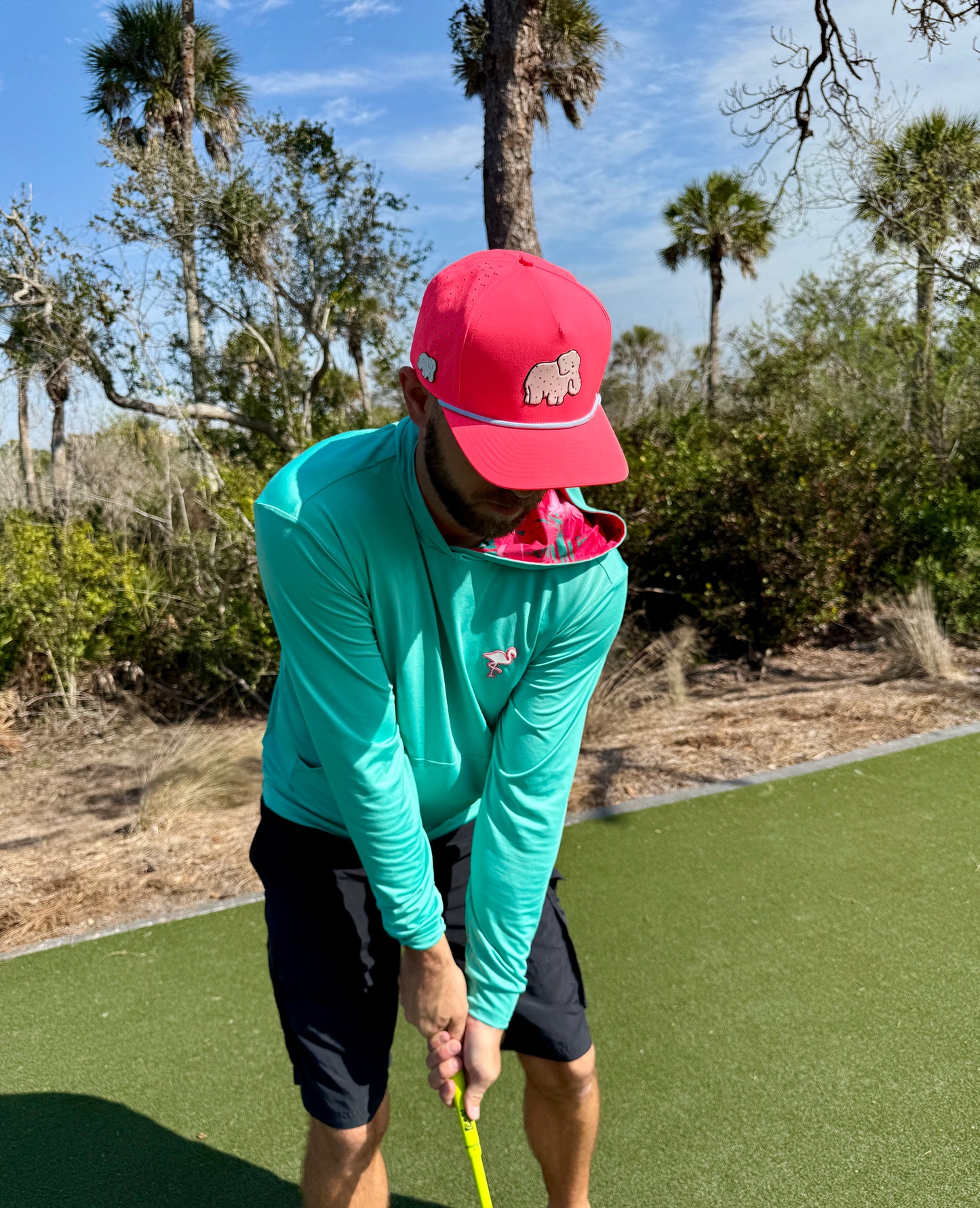 A person in a turquoise shirt and an Elephant Cookie Pink Frosted Elephant Golf Hat with a built-in magnetic ball marker swings a golf club on a green with palm trees and dry vegetation under a blue sky.