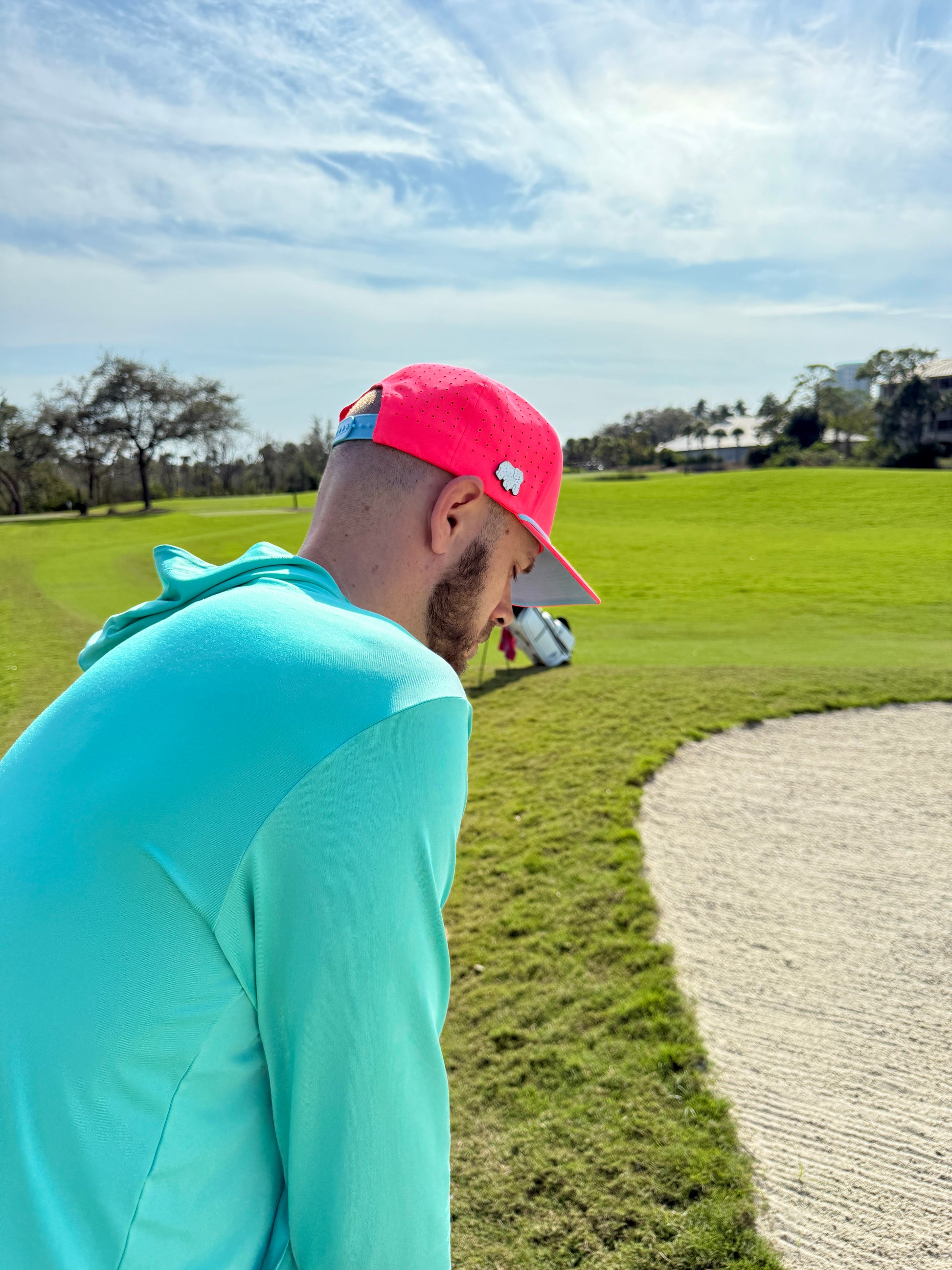 A man in a teal hoodie and the Frosted Elephant Golf Hat—an adjustable snapback cap with a magnetic ball marker holder, included marker, and Elephant Cookie Pink design—stands by a sand trap under a blue sky on a golf course.