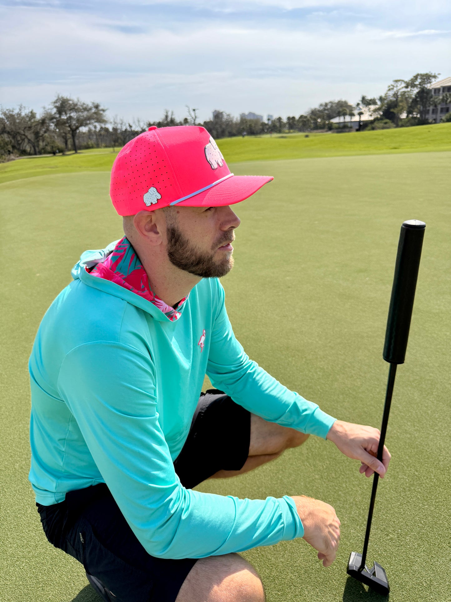 A man in an Elephant Cookie Pink Frosted Elephant Golf Hat with a built-in magnetic ball marker holder kneels on the green, putter in hand, wearing an aqua blue hoodie, under a sunny sky with trees and buildings behind him.