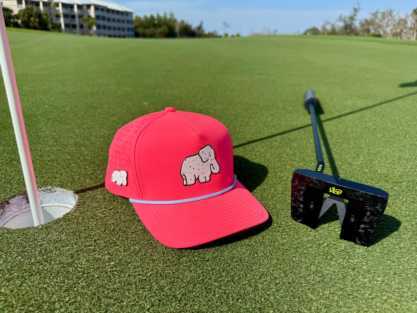 The Frosted Elephant Golf Hat in Elephant Cookie Pink features an embroidered white elephant, built-in magnetic ball marker holder with marker, and adjustable snapback. Perfect for golfers, it stands out on the green beside your putter and flagstick.
