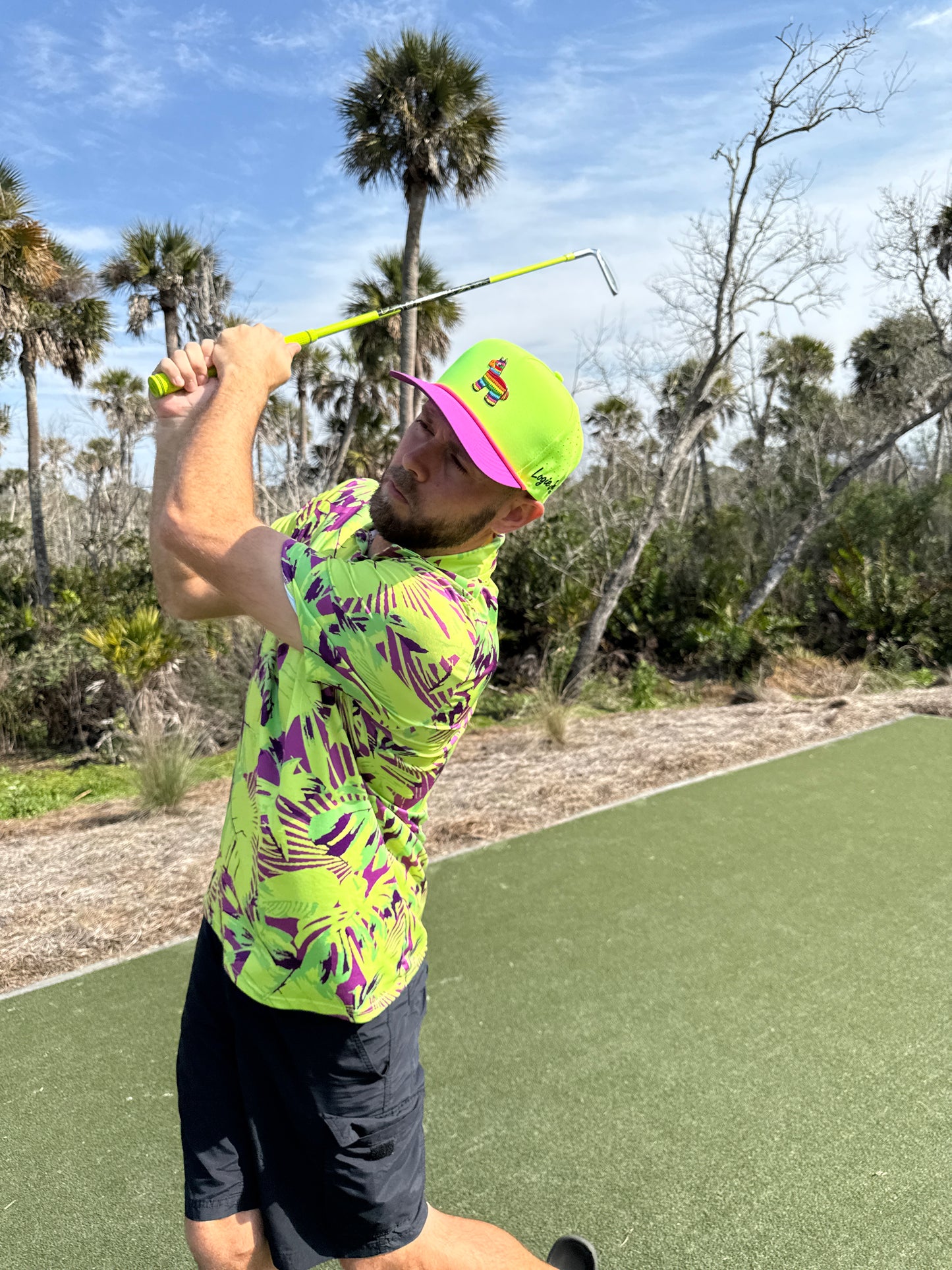 A man in a tropical shirt and the "I'd Hit That Piñata Golf Hat" with llama design swings a club on a sunny course. The snapback cap features a built-in magnetic ball marker holder, included marker, and is fully adjustable.