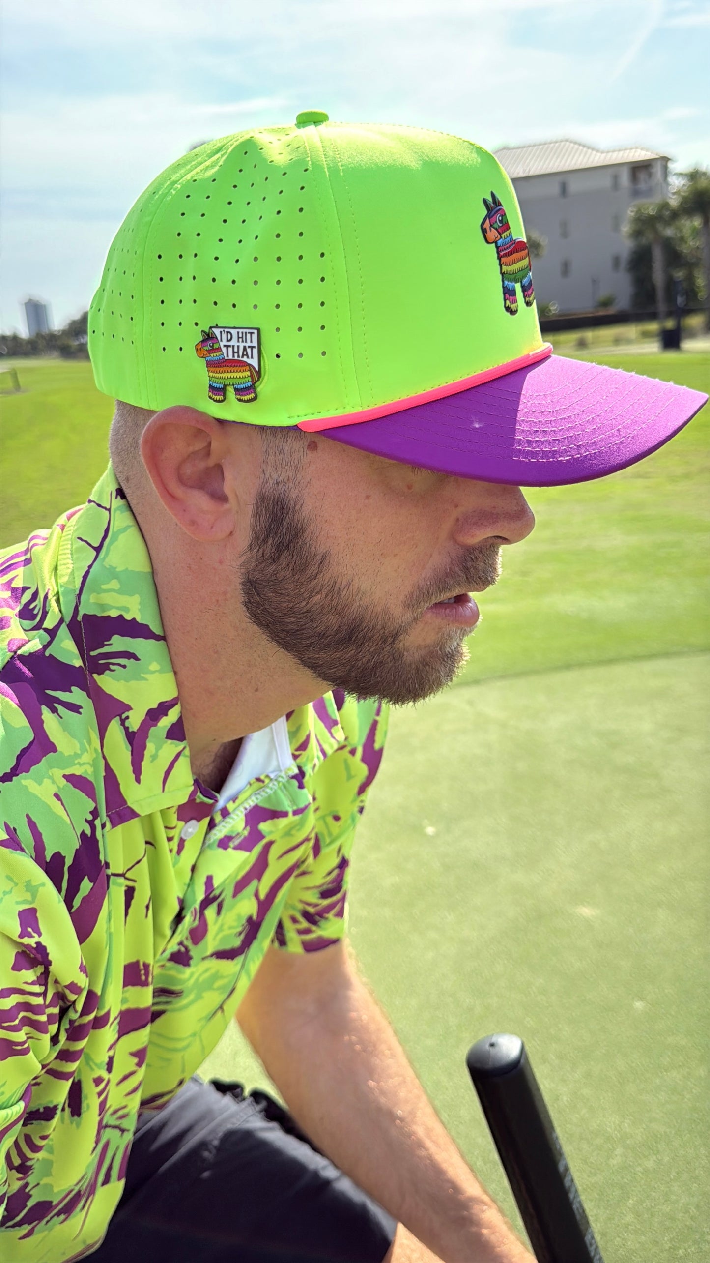A man wears the “I'd Hit That Piñata Golf Hat”—a llama multicolor snapback with a built-in magnetic ball marker holder—while golfing. He attaches his marker to the hat for easy access, standing on a grassy course under a blue sky.