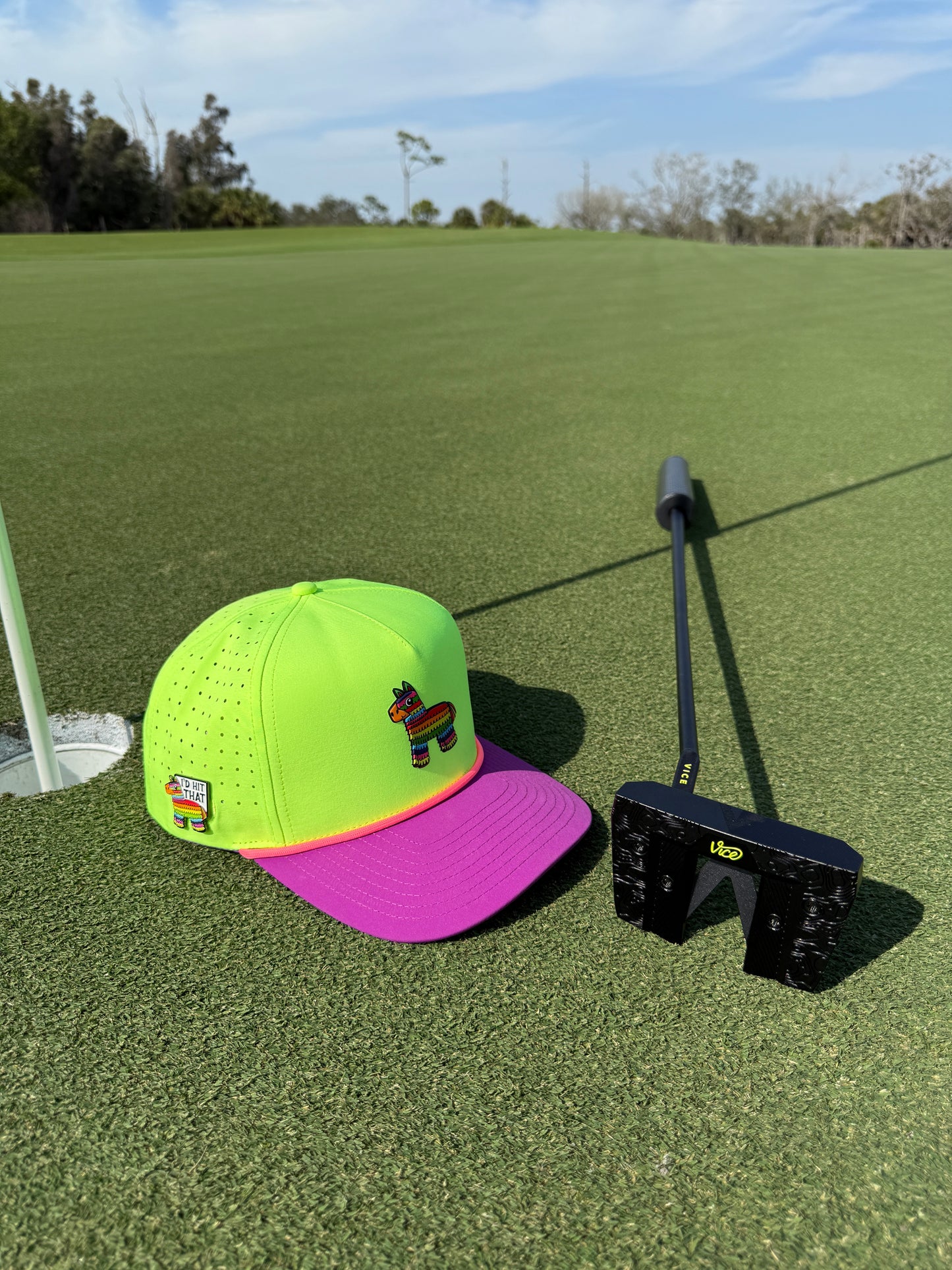 The I'd Hit That Piñata Golf Hat, a multicolor llama snapback with a built-in magnetic ball marker holder and included marker, sits on a putting green next to a black putter and marker near the hole on a sunny golf course.