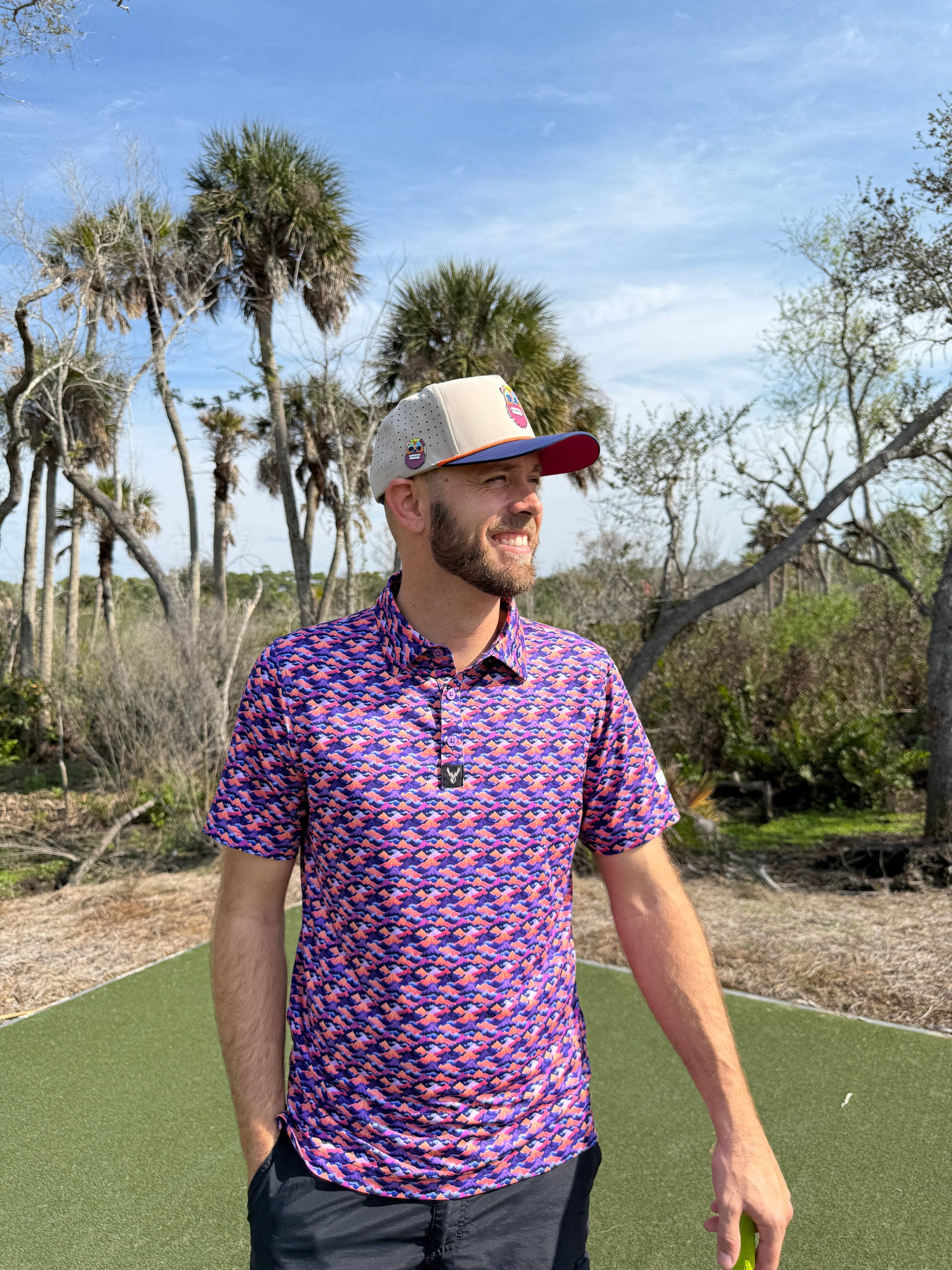A bearded man wearing a colorful polo and the Tiki Bear Golf Hat—an adjustable multicolor snapback cap with a built-in magnetic ball marker holder—stands smiling on a green mat outdoors, palm trees behind him under a sunny sky.