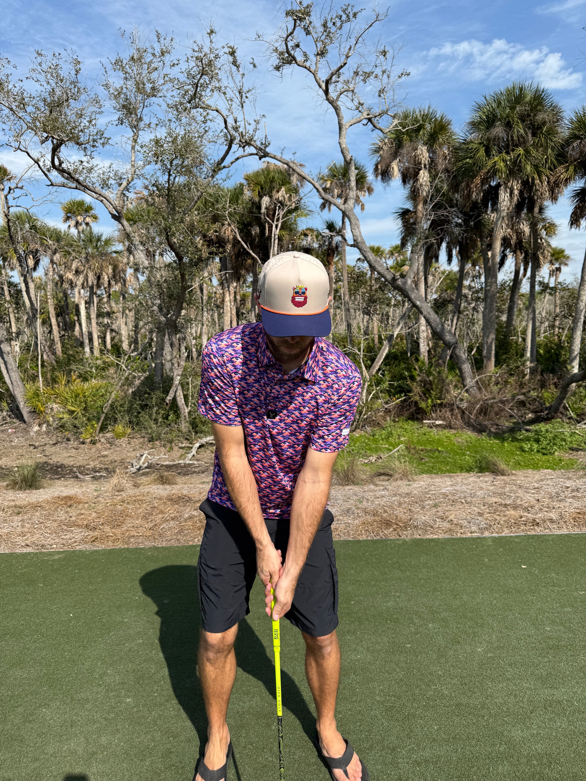 A man in a colorful shirt, shorts, and the Tiki Bear Golf Hat—an adjustable multicolor snapback with built-in magnetic ball marker holder—stands on artificial turf outdoors, preparing to swing a golf club amid palm trees and blue sky.