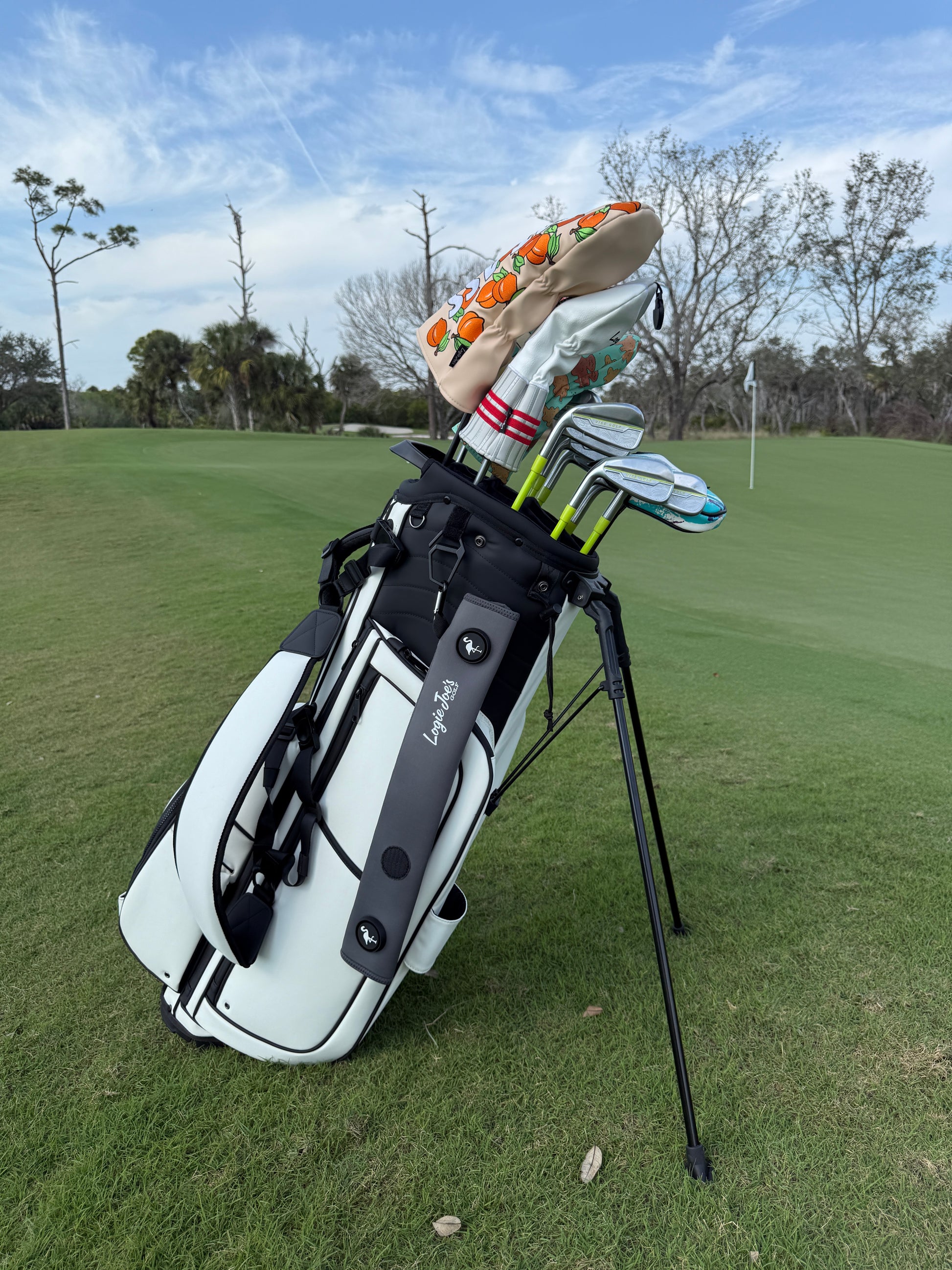 A white golf bag with clubs stands on green grass near a green. Pops of orange come from accessories, including a gray neoprene magnetic golf cart strap sleeve (18.5x3.5 in) with dual magnets, glove holder, and carabiner. Trees and flagstick under cloudy sky.