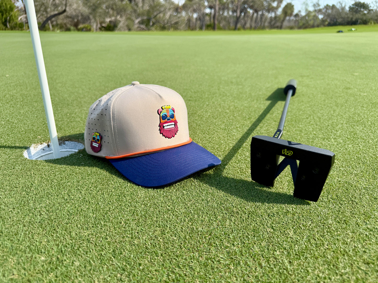 A golf putter and a Tiki Bear Golf Hat Snapback Cap with a colorful cartoon face, built-in magnetic ball marker holder, and included marker rest on the green near the hole and flag, with trees in the background.