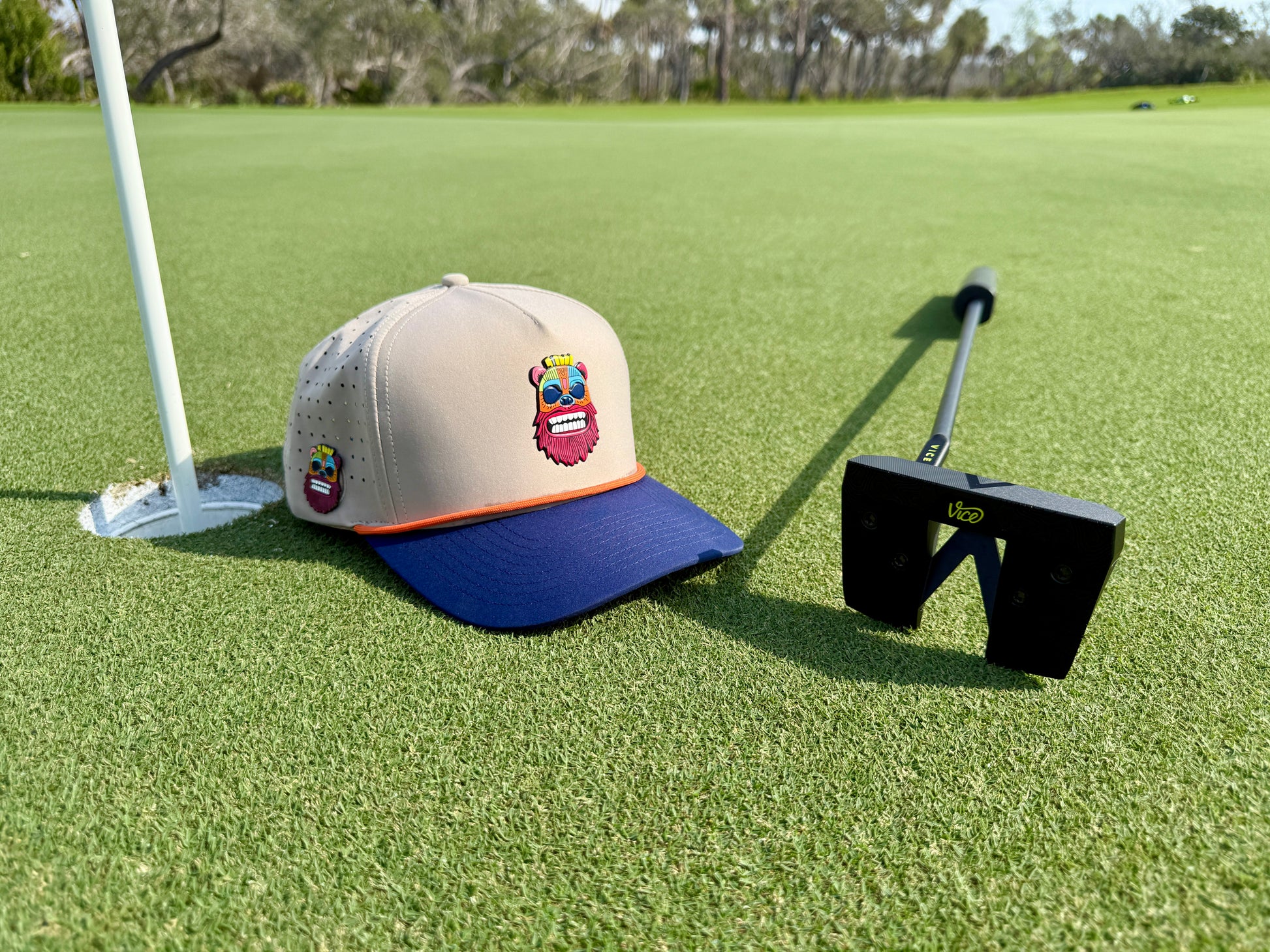 A golf putter and a Tiki Bear Golf Hat Snapback Cap with a colorful cartoon face, built-in magnetic ball marker holder, and included marker rest on the green near the hole and flag, with trees in the background.