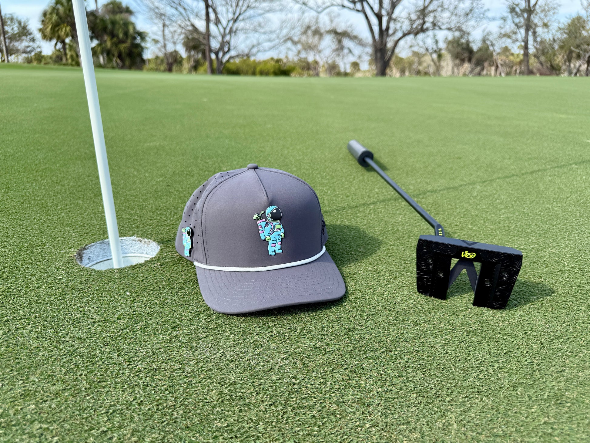 The Astronaut Walker Golf Hat | Snapback Cap in gray, featuring a built-in magnetic ball marker holder and marker, rests near a golf hole and putter on green grass, with trees blurred in the background on a sunny day.
