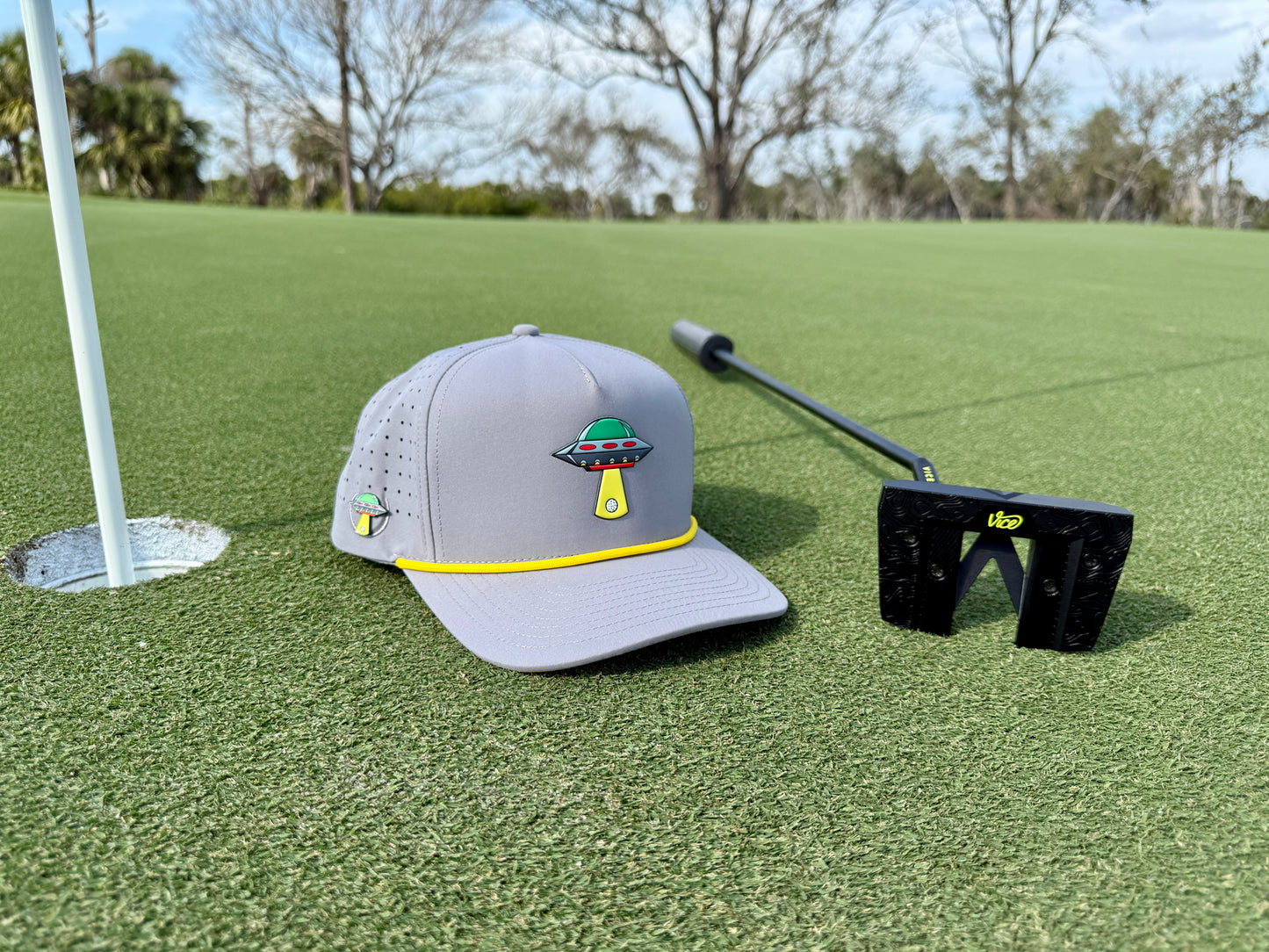 An Alien Gray UFO Golf Hat (Snapback Cap) with built-in magnetic ball marker holder and included marker, a putter, and a ball marker rest on the green near the hole, surrounded by trees under a partly cloudy sky.