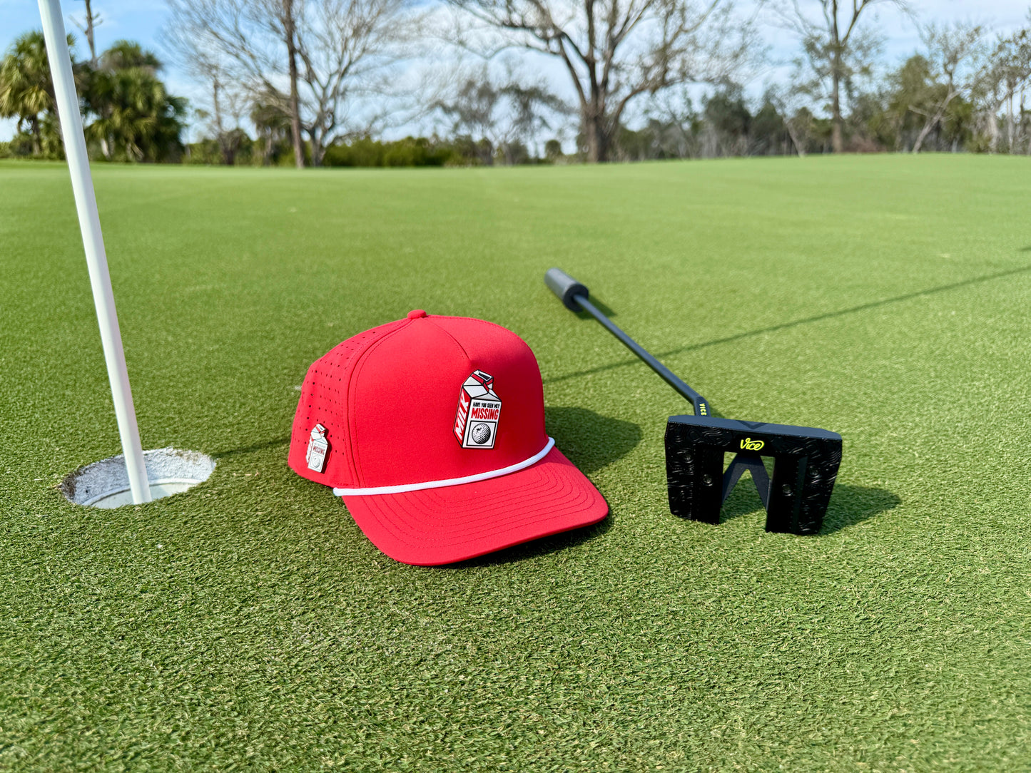 A Missing Milk Golf Hat—an adjustable red snapback cap with a built-in magnetic ball marker holder and marker—rests beside a black putter on the green, near the hole with flagstick, framed by trees and sky.