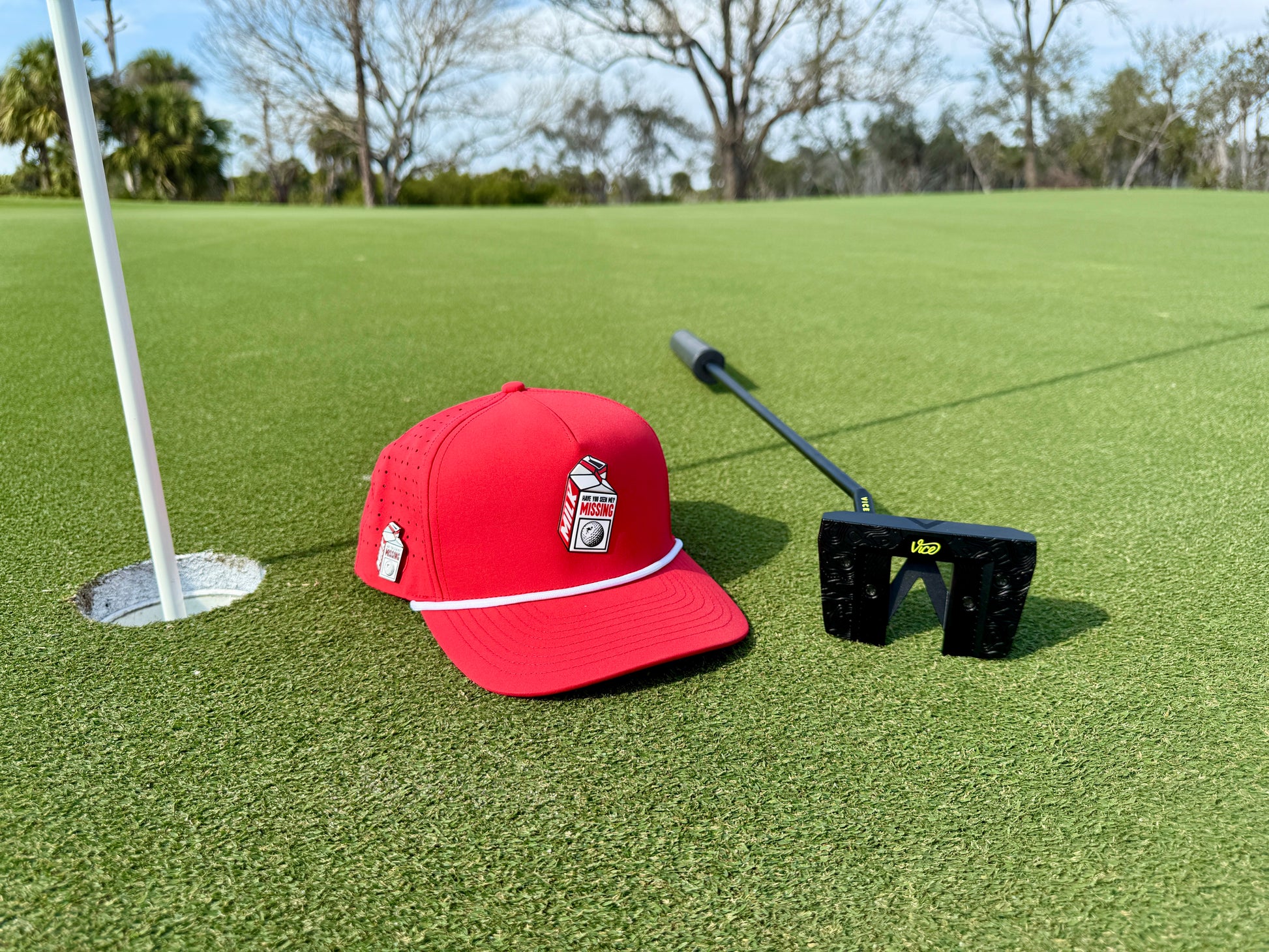 A Missing Milk Golf Hat—an adjustable red snapback cap with a built-in magnetic ball marker holder and marker—rests beside a black putter on the green, near the hole with flagstick, framed by trees and sky.