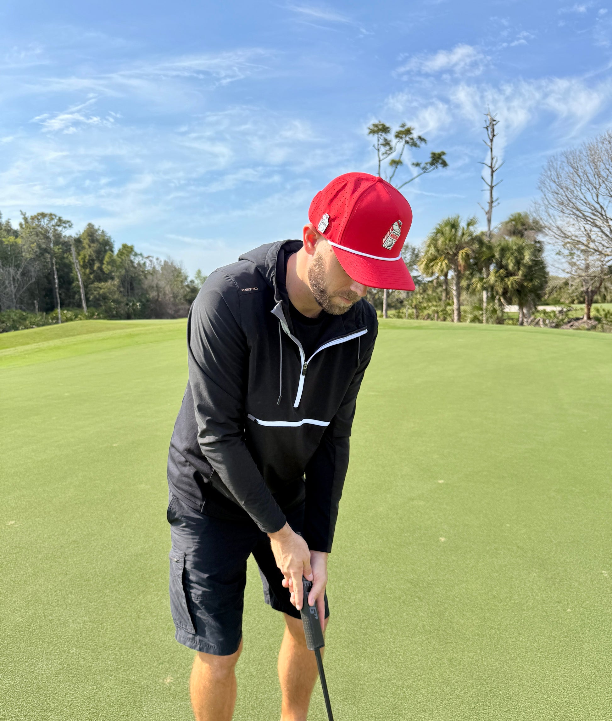 A man in an adjustable red Missing Milk Golf Hat with built-in magnetic ball marker focuses on putting a golf ball on a green course under a blue sky, wearing a black jacket and shorts, trees in the background.