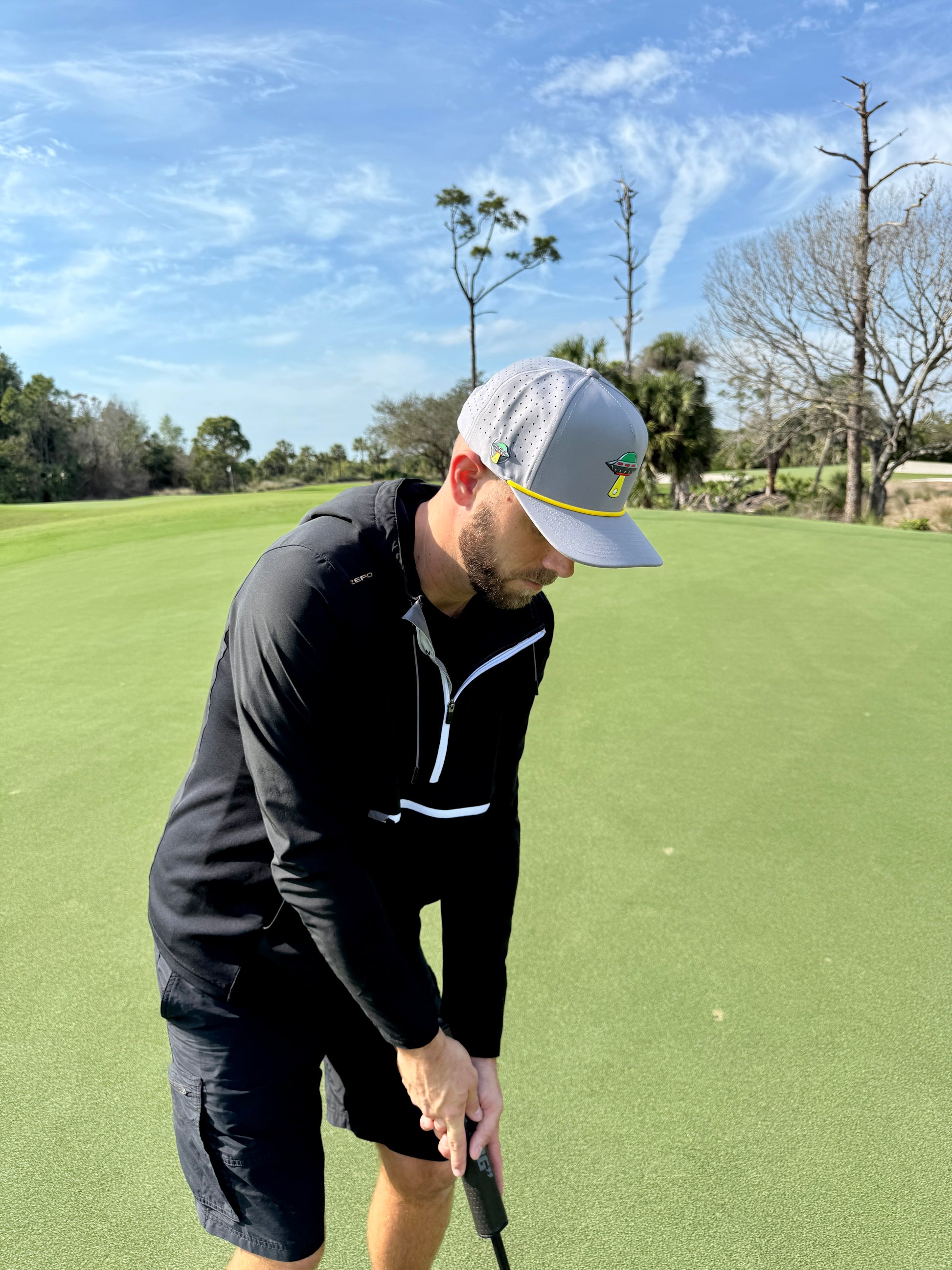 On a sunny golf course, a man in the UFO Golf Hat—Alien Gray snapback with built-in magnetic ball marker holder—prepares to putt, staying focused thanks to this essential adjustable cap.
