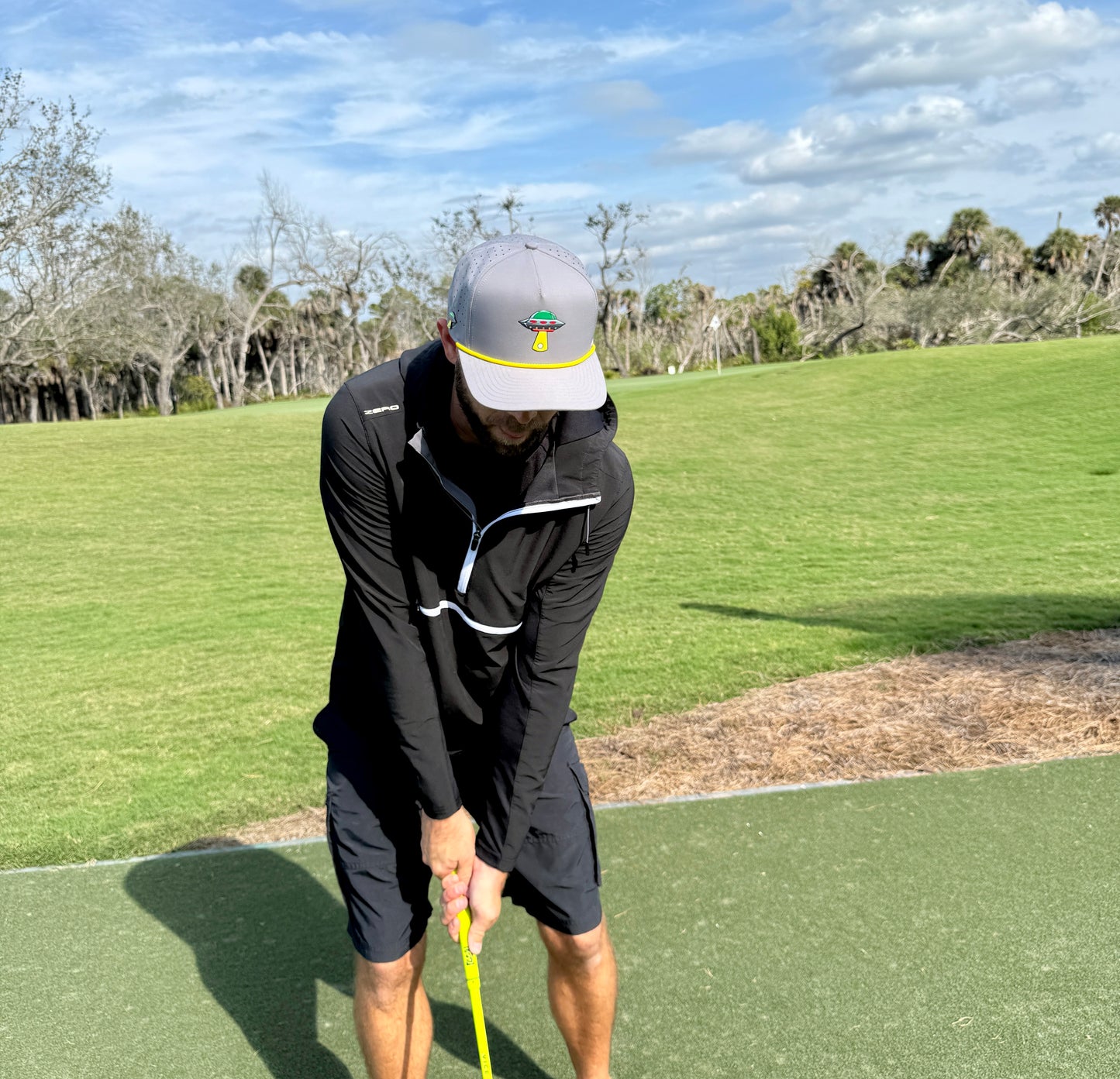 A man in an Alien Gray UFO Golf Hat with built-in magnetic ball marker holder and marker, adjustable snapback cap, practices putting on a green under a partly cloudy sky, surrounded by trees and grass.