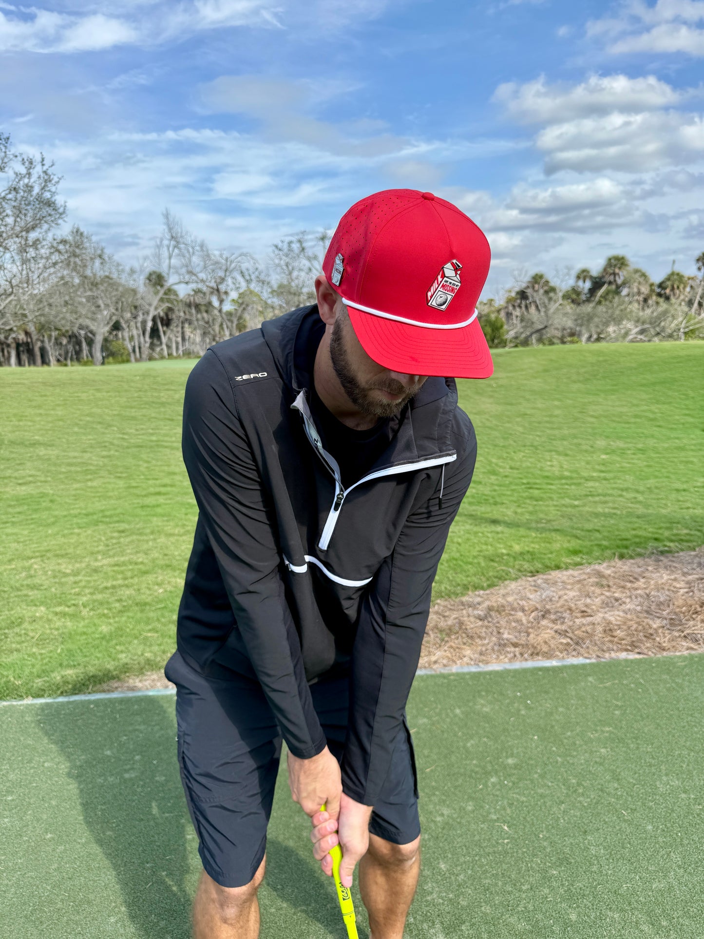 A man in a Missing Milk Golf Hat—an adjustable red snapback cap with a magnetic ball marker holder—is on the golf course in black gear, holding a yellow club and preparing to take a shot under partly cloudy skies.