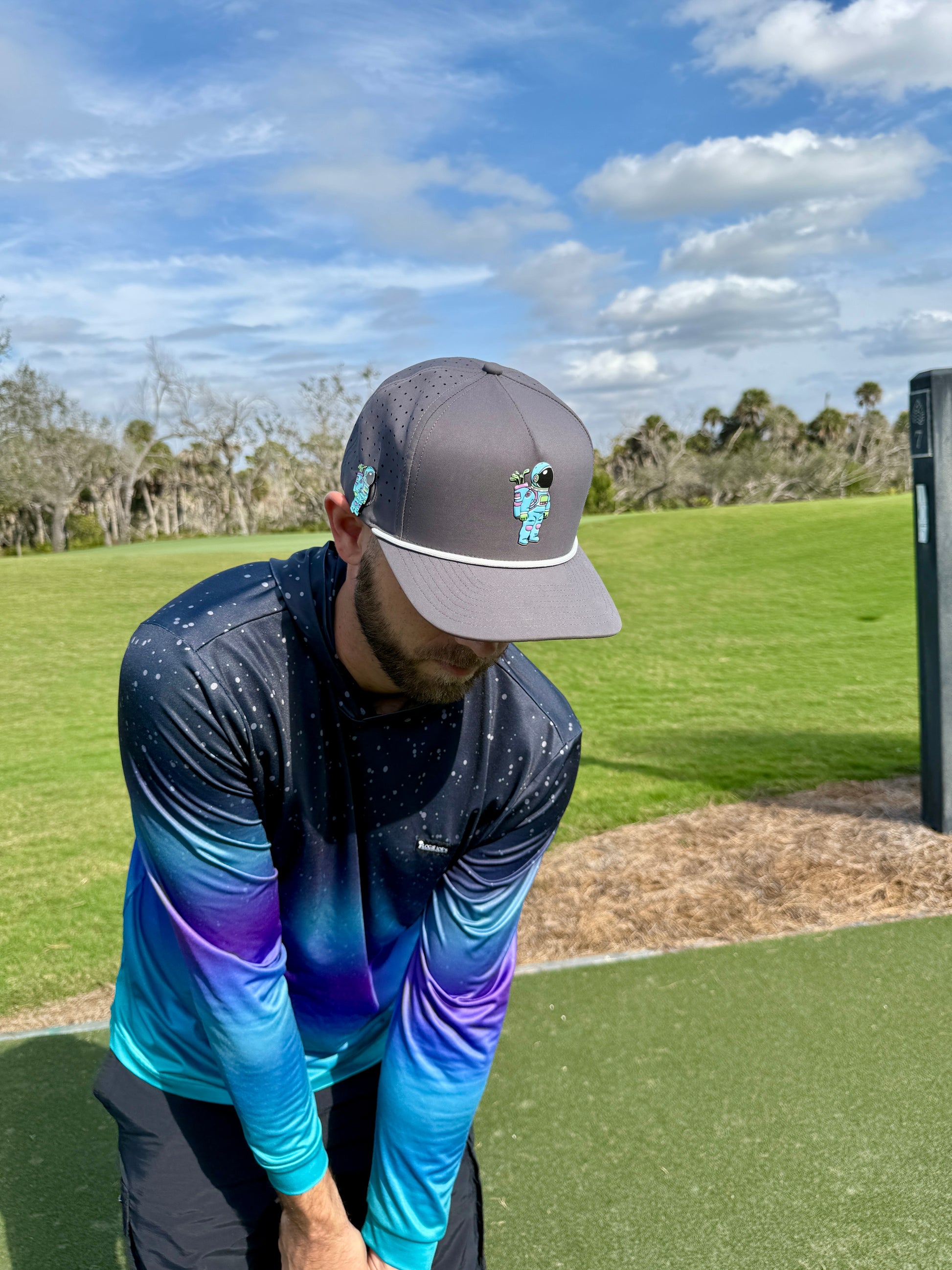 A man in an Astronaut Walker Golf Hat—an adjustable gray snapback cap with a built-in magnetic ball marker holder—and a blue-purple gradient long-sleeve shirt prepares to putt on a golf course, trees and clouds in the background.