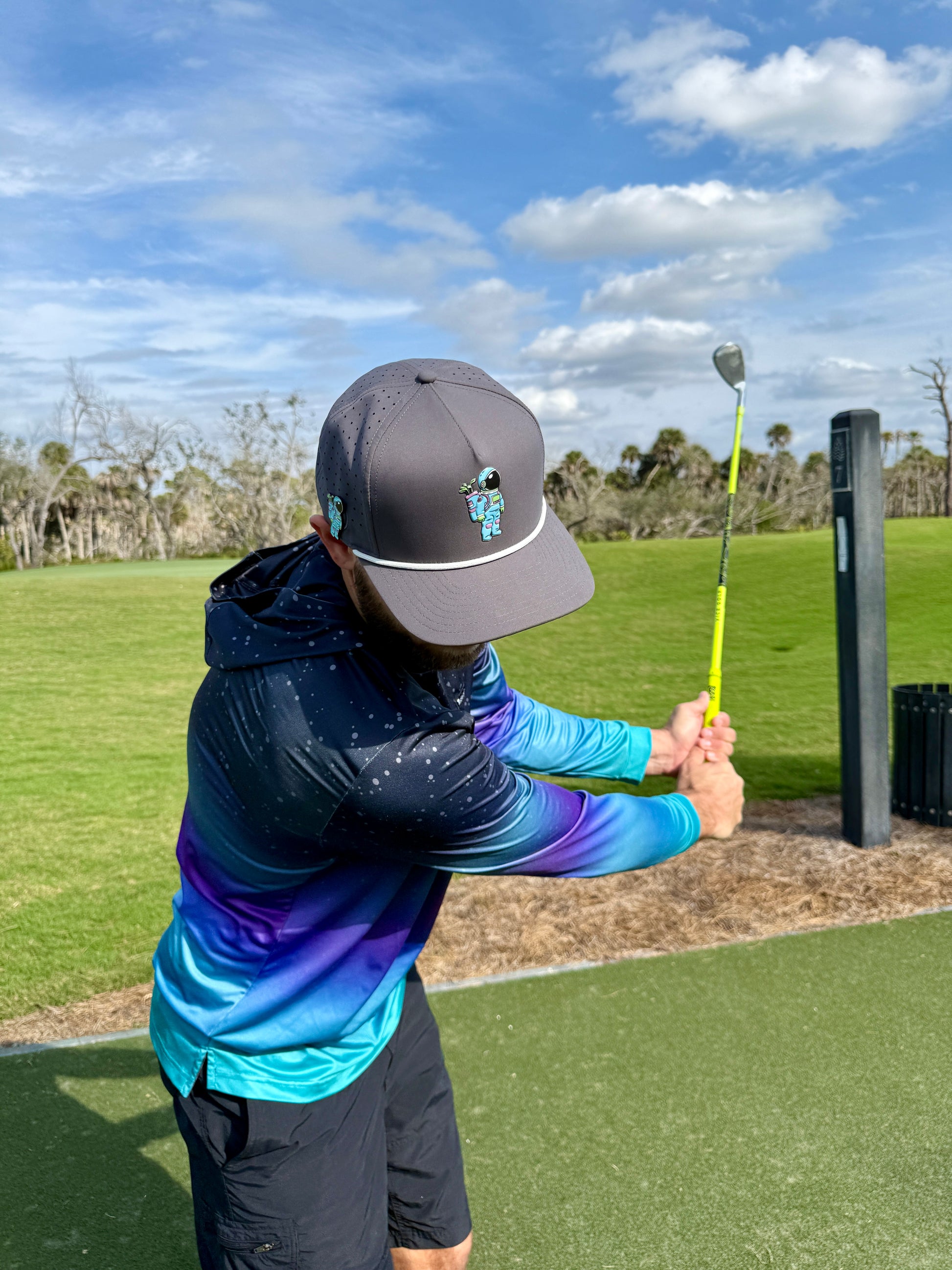 A person in a colorful blue and purple jacket and gray Astronaut Walker Golf Hat with magnetic ball marker holds a yellow golf club, preparing to swing on a grassy golf course under a partly cloudy sky.
