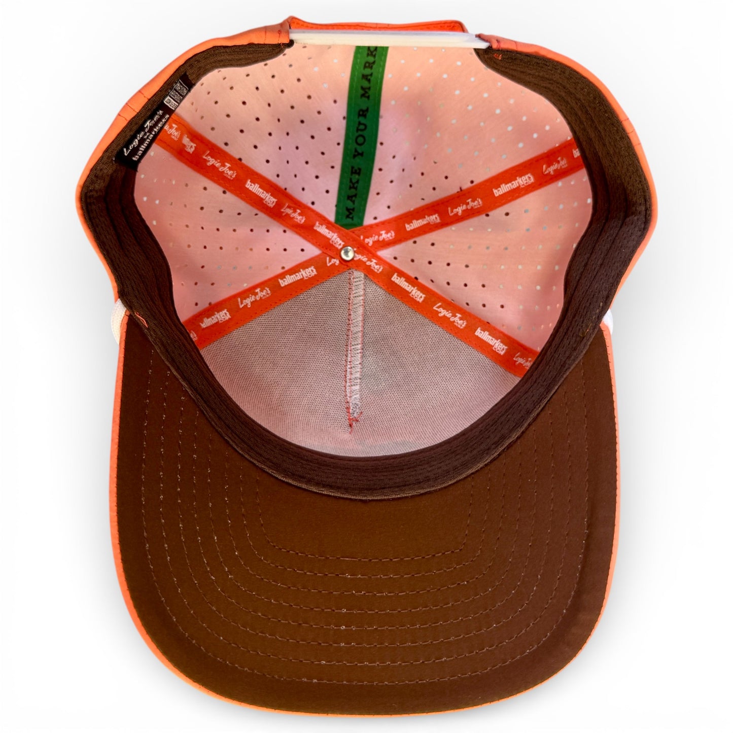 A look inside the Just Peachy Golf Hat reveals orange, brown, and green lining—the green says “MAKE YOUR MARK.” This adjustable peach snapback features perforated pink fabric, a brown brim, and a built-in magnetic ball marker holder with marker.