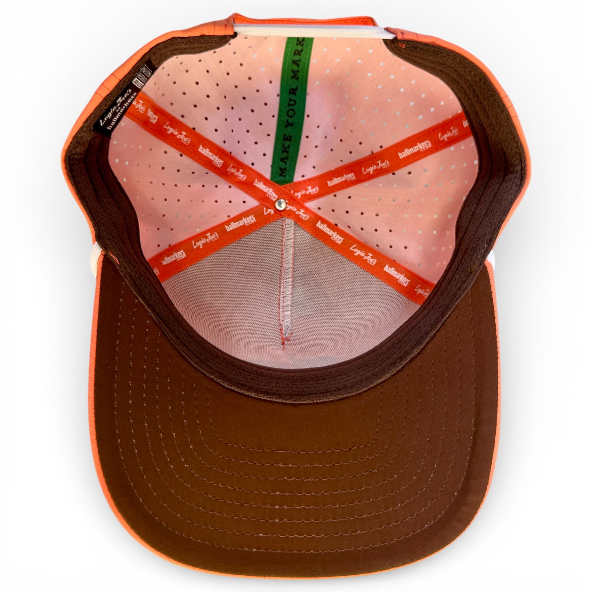A look inside the Just Peachy Golf Hat reveals orange, brown, and green lining—the green says “MAKE YOUR MARK.” This adjustable peach snapback features perforated pink fabric, a brown brim, and a built-in magnetic ball marker holder with marker.