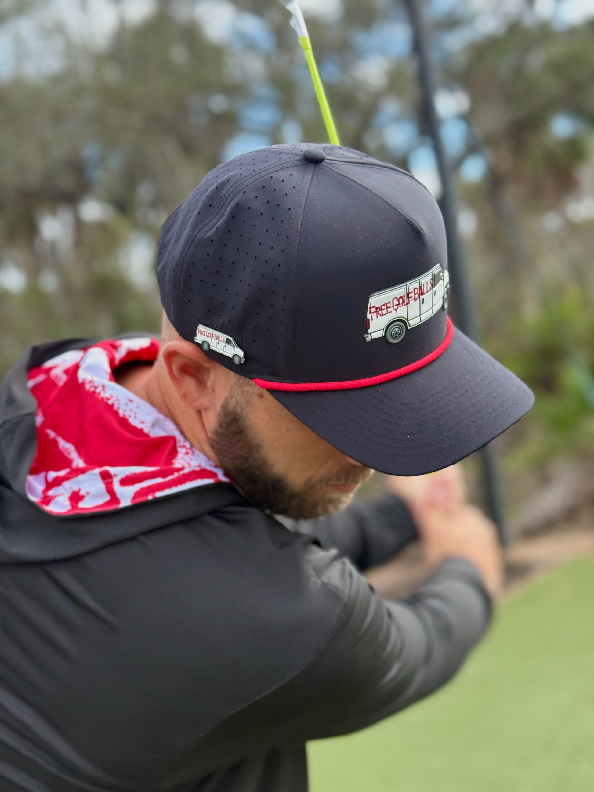 A man swings a golf club outdoors, wearing the Free Golf Balls Van Golf Hat—an adjustable black snapback cap with built-in magnetic ball marker holder and included marker. Trees and greenery are visible in the background.