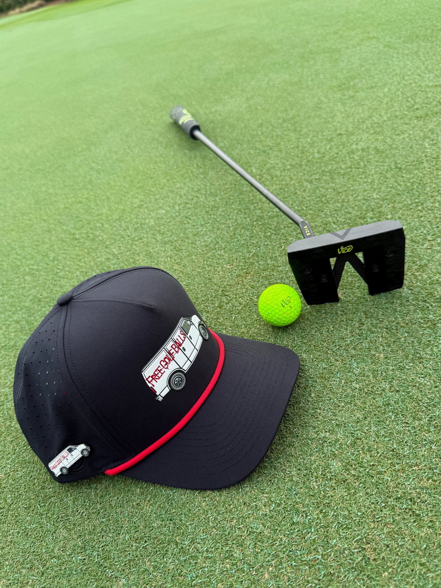 A Free Golf Balls Van Golf Hat with built-in magnetic ball marker holder (marker included) and a cartoon bus patch, plus a yellow-green golf ball and black putter, rest on a putting green. The adjustable black snapback cap also features an enamel bus pin.