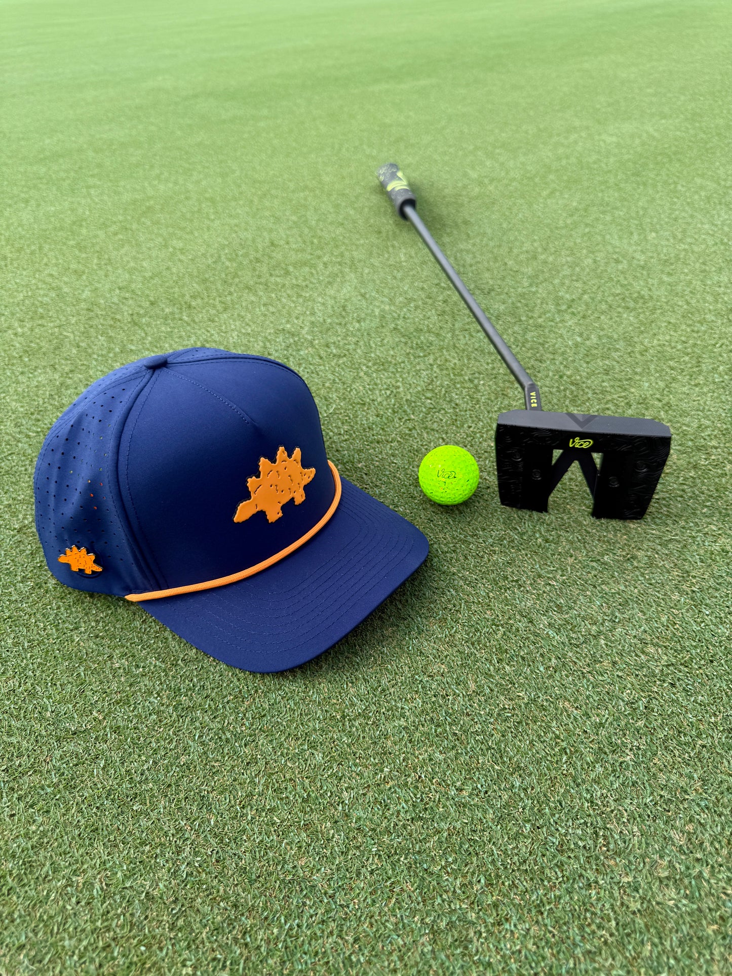 A blue and orange Dino Nugget Golf Hat with adjustable snapback and built-in magnetic ball marker holder, a bright green golf ball with marker, and a black putter on neatly trimmed green.