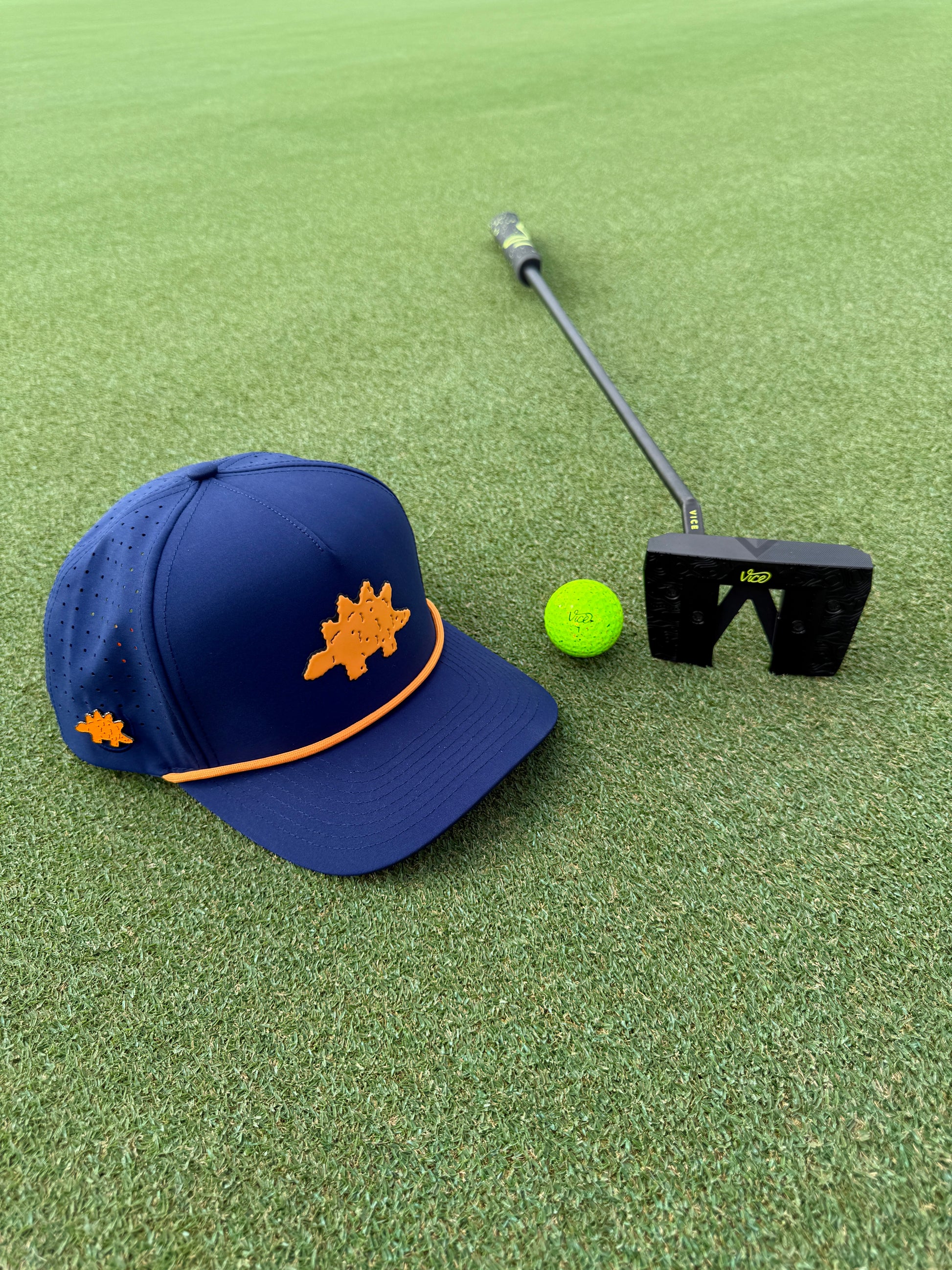 A blue and orange Dino Nugget Golf Hat with adjustable snapback and built-in magnetic ball marker holder, a bright green golf ball with marker, and a black putter on neatly trimmed green.
