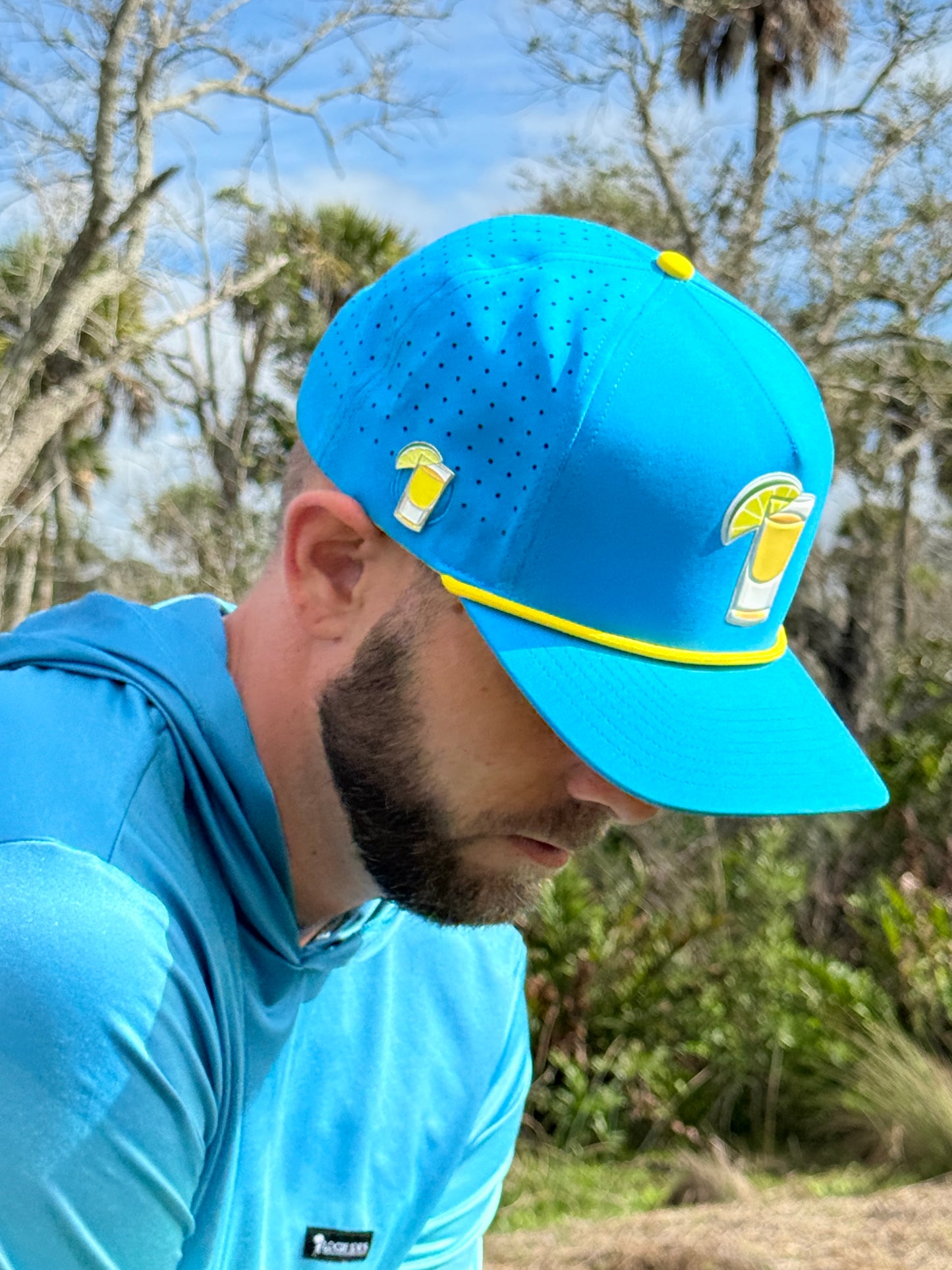 A man in a bright Tequila and Lime Golf Hat with built-in magnetic ball marker and matching shirt, featuring a lemon drink patch, looks down outdoors surrounded by trees and greenery on a sunny day.