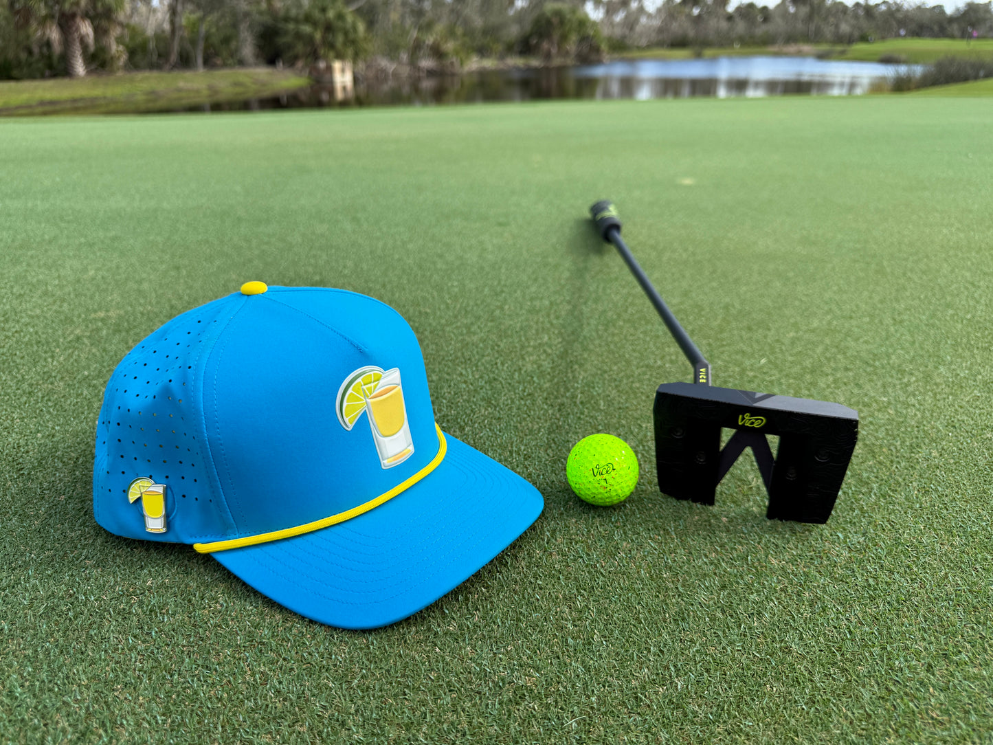 A Tequila and Lime Golf Hat snapback with built-in magnetic ball marker holder and included marker, a neon yellow golf ball, and a black putter rest on the green course by water, framed by trees and blue sky.