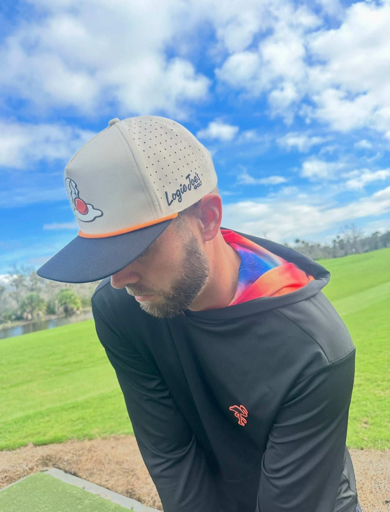 A man wearing the Breakfast Ball Golf Hat in Mulligan Tan—a snapback cap with adjustable fit, built-in magnetic ball marker holder, and included marker—paired with a black jacket.