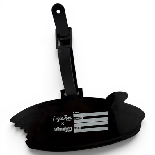 The Shark Bite Clip-On Tag is a black luggage tag shaped like a bitten surfboard, with fields for name, address, and phone. It features Logie Joes x ballmarkers in white and comes with a black strap—ideal as a golf bag accessory.