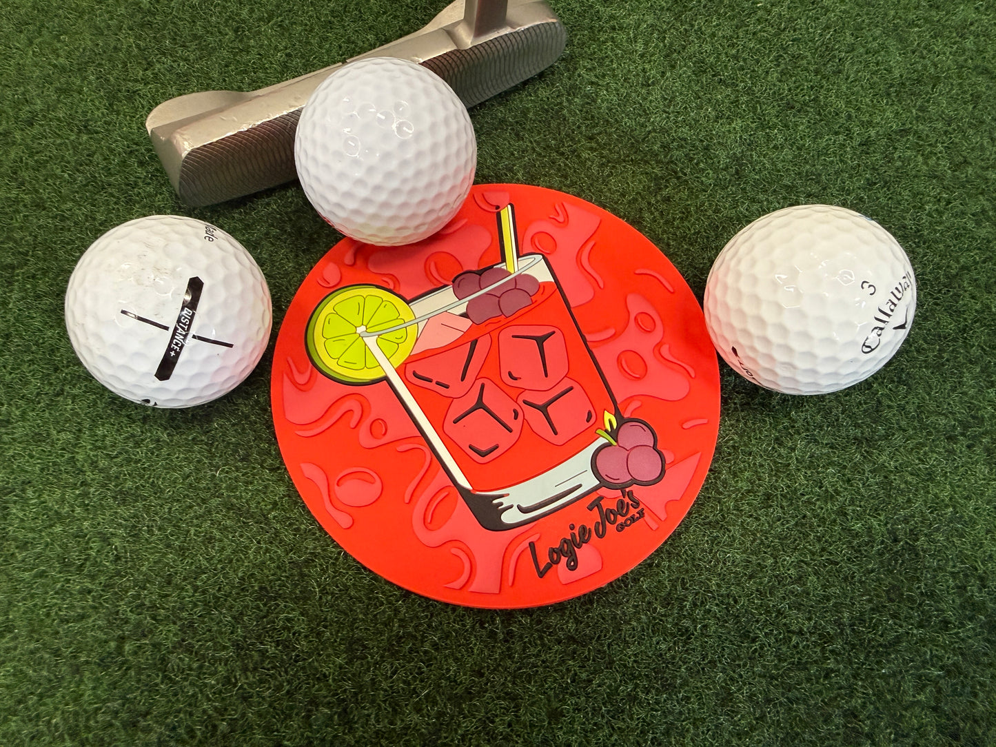 Three golf balls and a putter surround the Transfusion Golf Putting Disk, an indoor practice marker featuring a cocktail graphic, placed on artificial grass—a portable golf training aid and unique putting target.