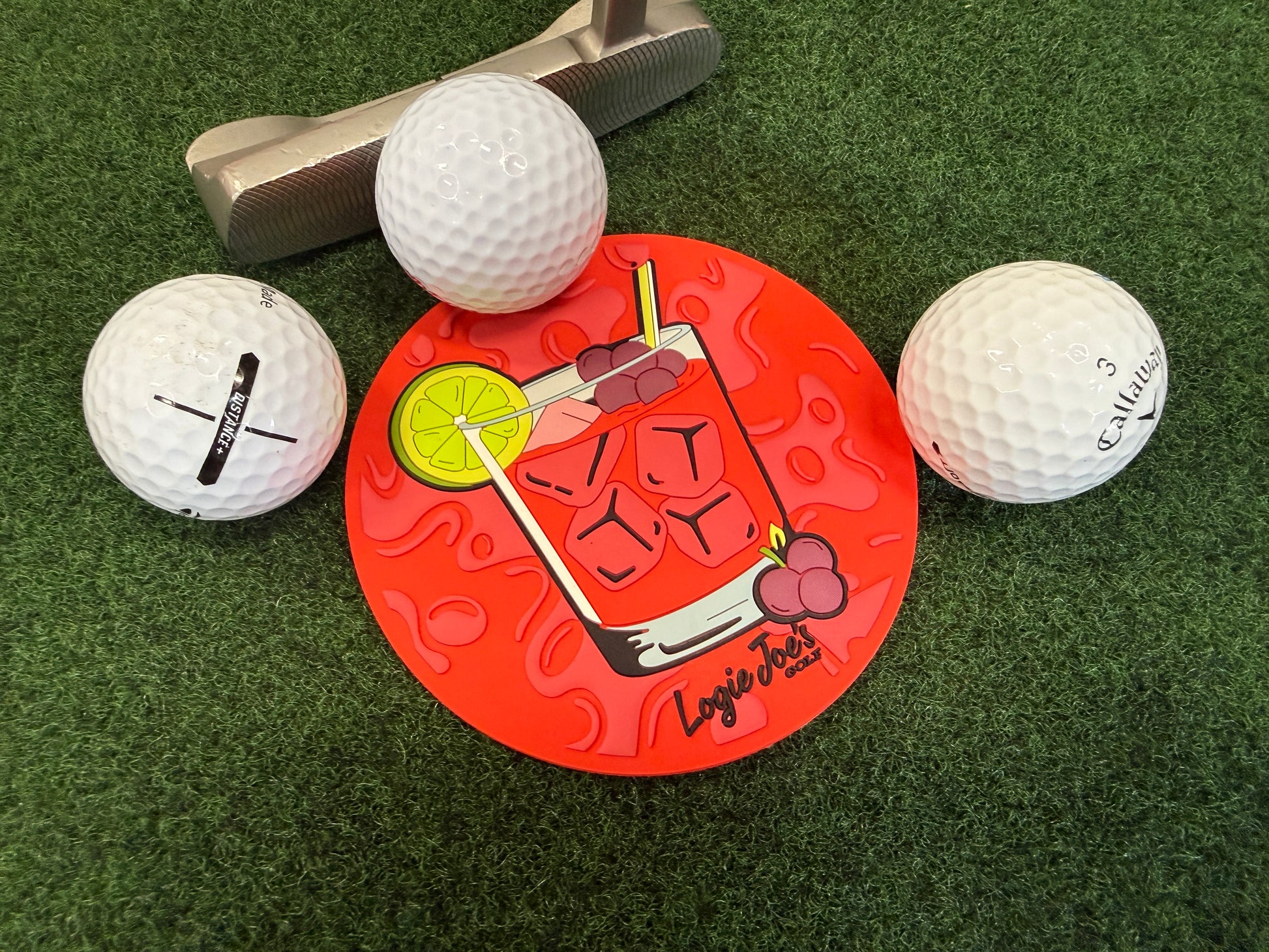 Three golf balls and a putter surround the Transfusion Golf Putting Disk, an indoor practice marker featuring a cocktail graphic, placed on artificial grass—a portable golf training aid and unique putting target.