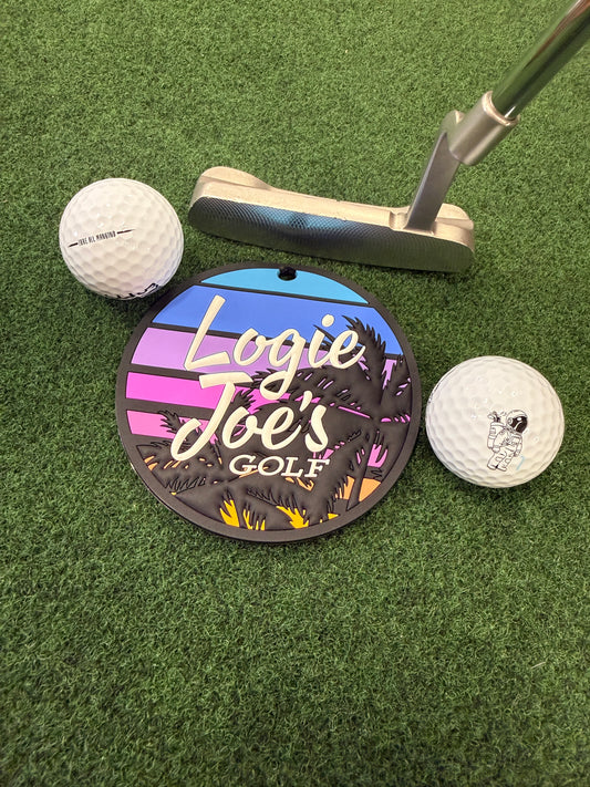A golf putter and two balls sit on green turf near a colorful Sunset Palms Golf Putting Disc, featuring palm trees and a sunset, with this portable indoor practice marker enhancing the putting experience.