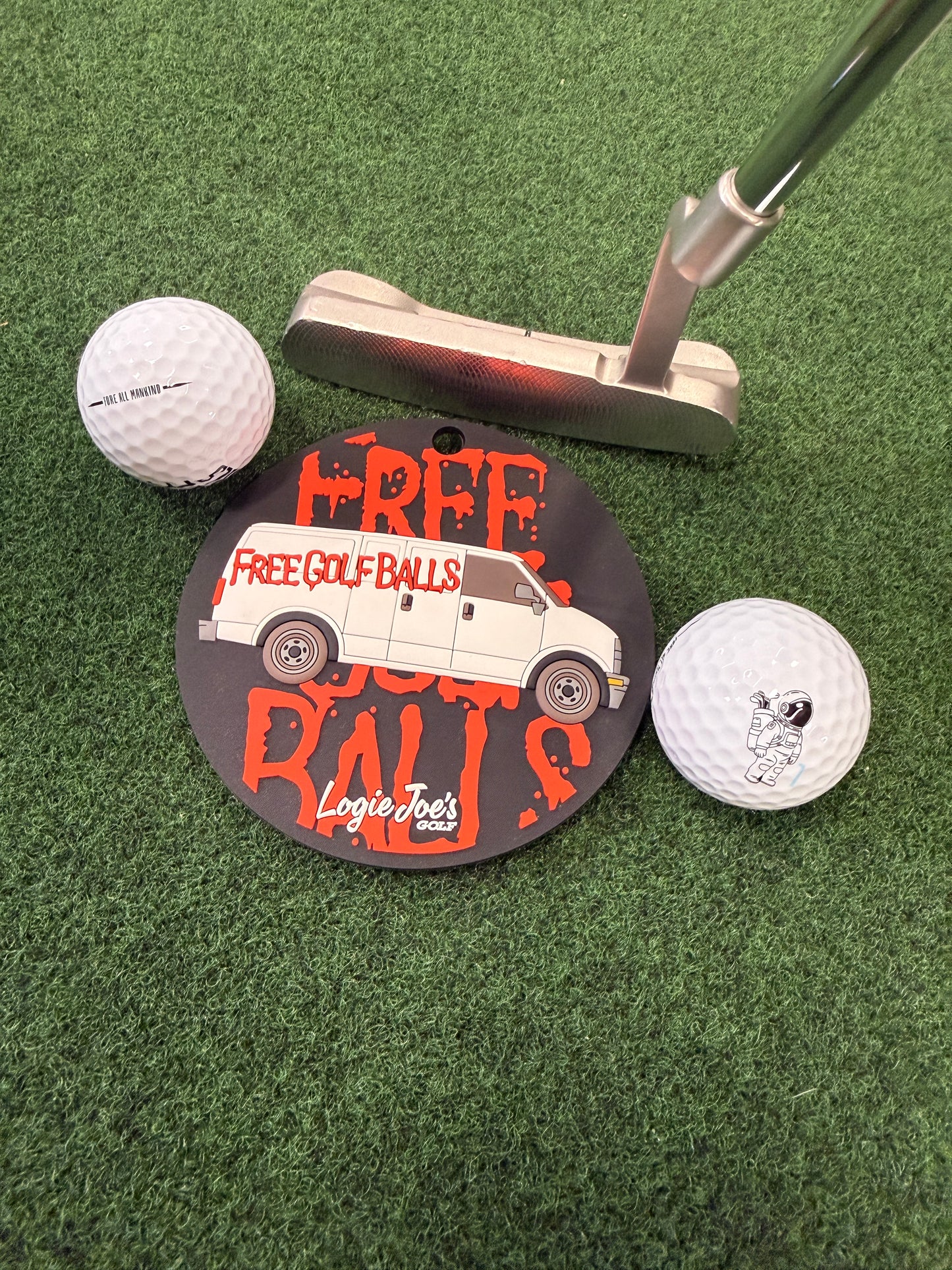 The Free Golf Balls Van Golf Putting Disc, with two balls and a circular "Free Golf Balls" sign featuring the Logie Joes logo, sits on green turf—an ideal indoor practice marker and portable golf training aid.
