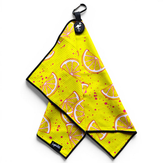 The Main Squeeze Golf Towel is a 24” x 16” yellow microfiber towel with lemon slice and pink splatter designs, black edging, a logo tag, and a magnetic clip plus carabiner for convenience on the course.