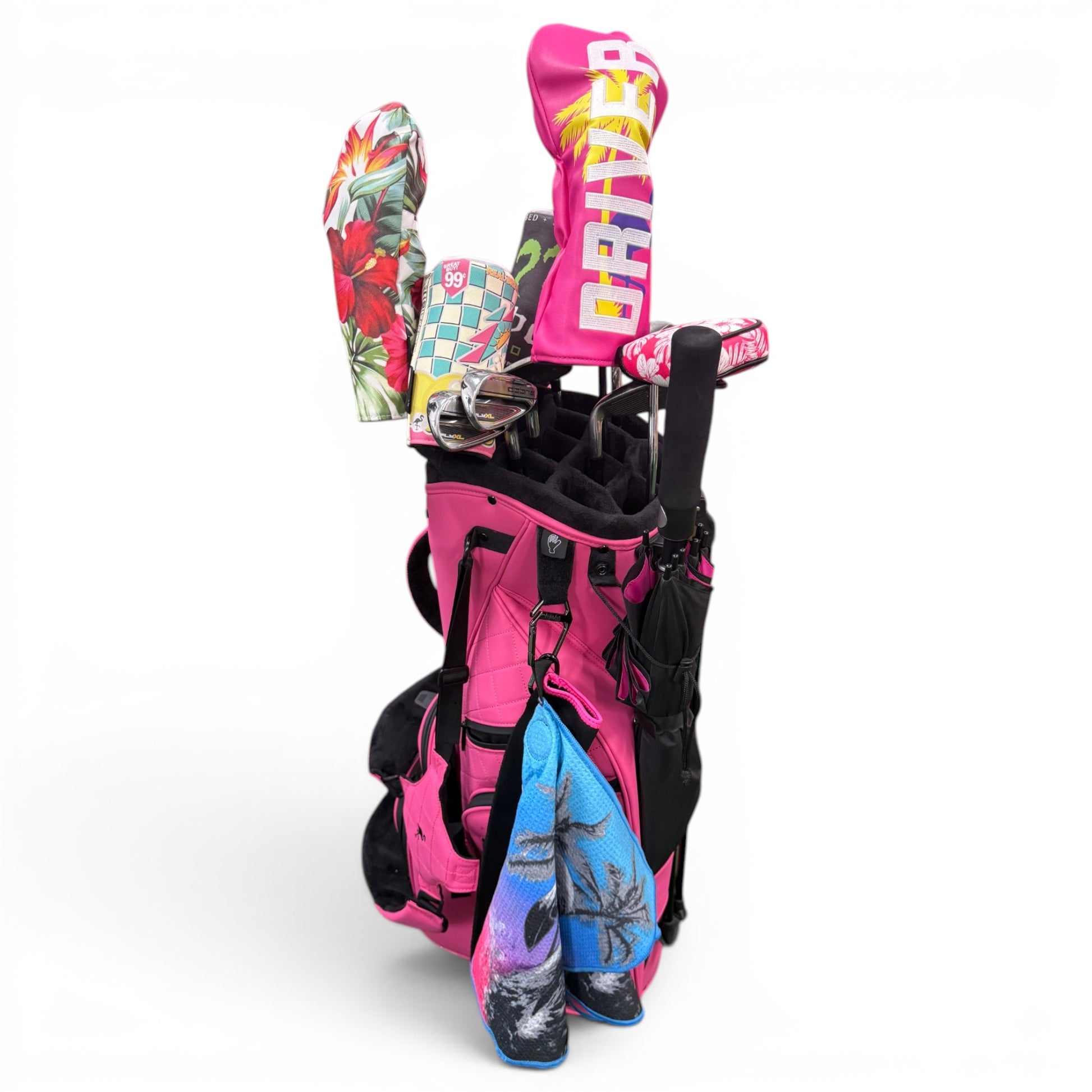 A bright pink golf bag holds clubs with colorful headcovers—one boldly labeled DRIVER. An oversized 60" Black & Pink Golf Umbrella provides rain protection, while two vibrant towels with tropical designs hang from the bag.