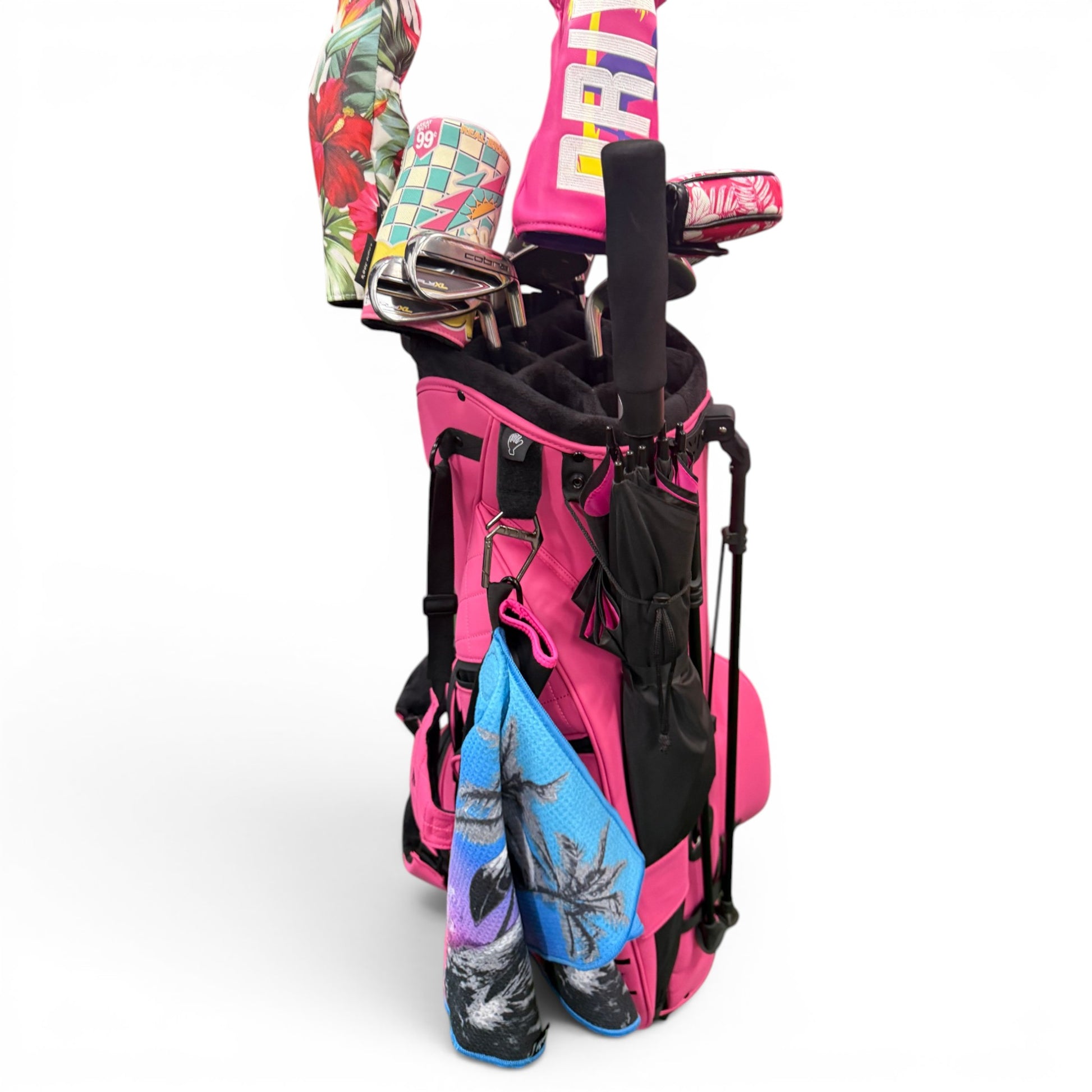 A bright pink golf bag with clubs sporting colorful headcovers (tropical and checkered) sits on a white background, alongside two palm tree towels and a 60" Golf Umbrella | Oversized Black & Pink Golf Umbrella for ample rain protection.