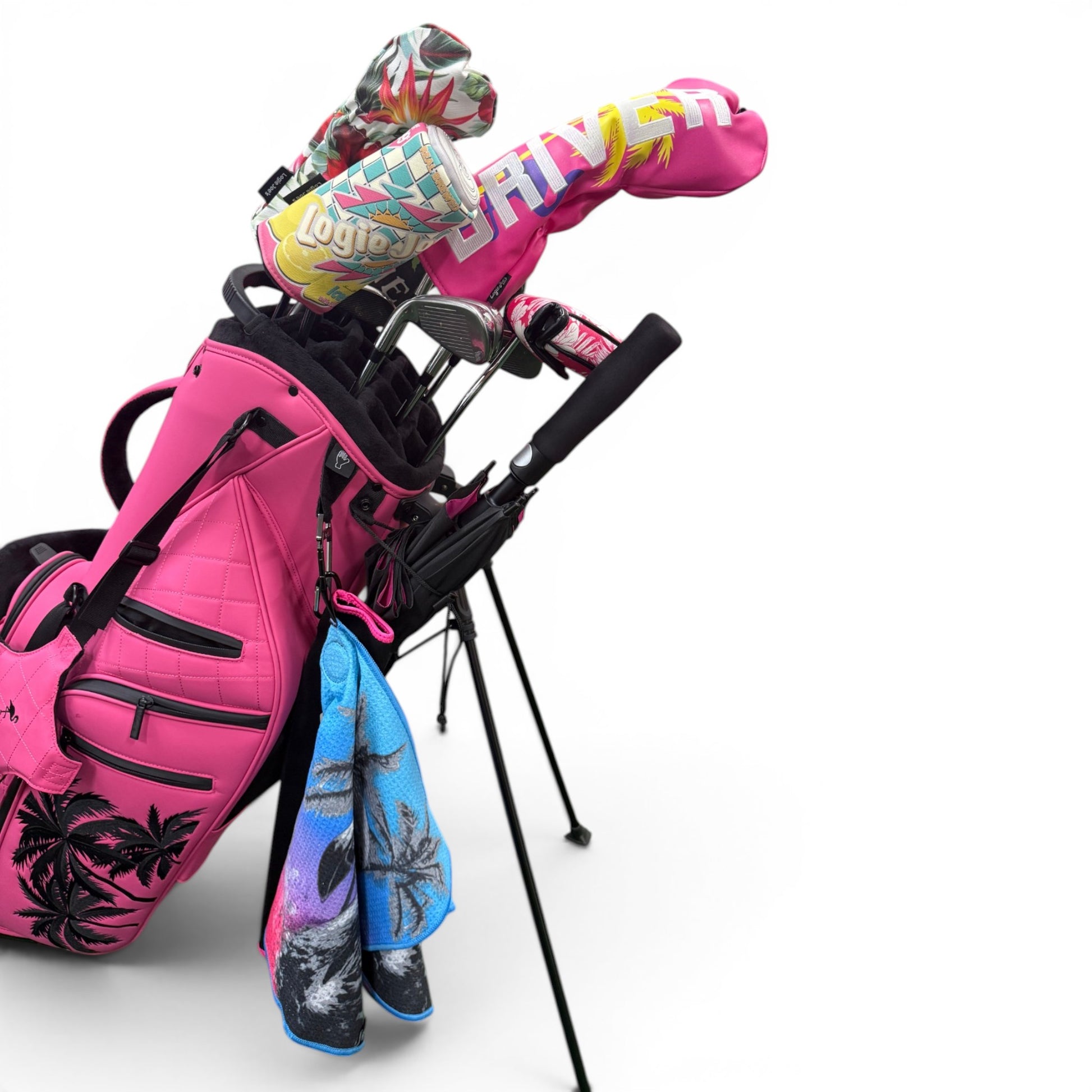 A bright pink golf bag with palm tree prints holds clubs with colorful headcovers. A blue and purple towel hangs from the side, while a 60" Golf Umbrella | Oversized Black & Pink Golf Umbrella offers rain protection. The bag stands on black legs.