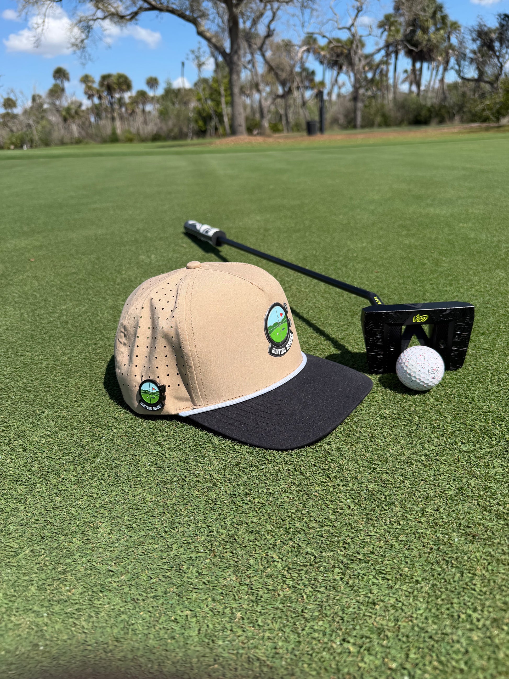 A tan and black Hunting Birdies Golf Hat with built-in magnetic ball marker holder and marker rests on the putting green by a golf ball and putter, trees and blue sky behind, showcasing this stylish adjustable snapback cap.
